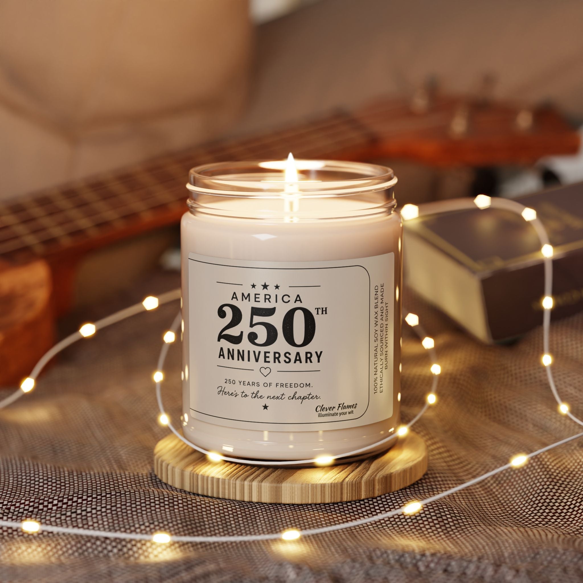 The Printify America 250th Anniversary Scented Soy Candle, 9oz with a patriotic label glows in its glass jar on a wooden coaster, surrounded by string lights—a charming Semiquincentennial keepsake beside a guitar and book.