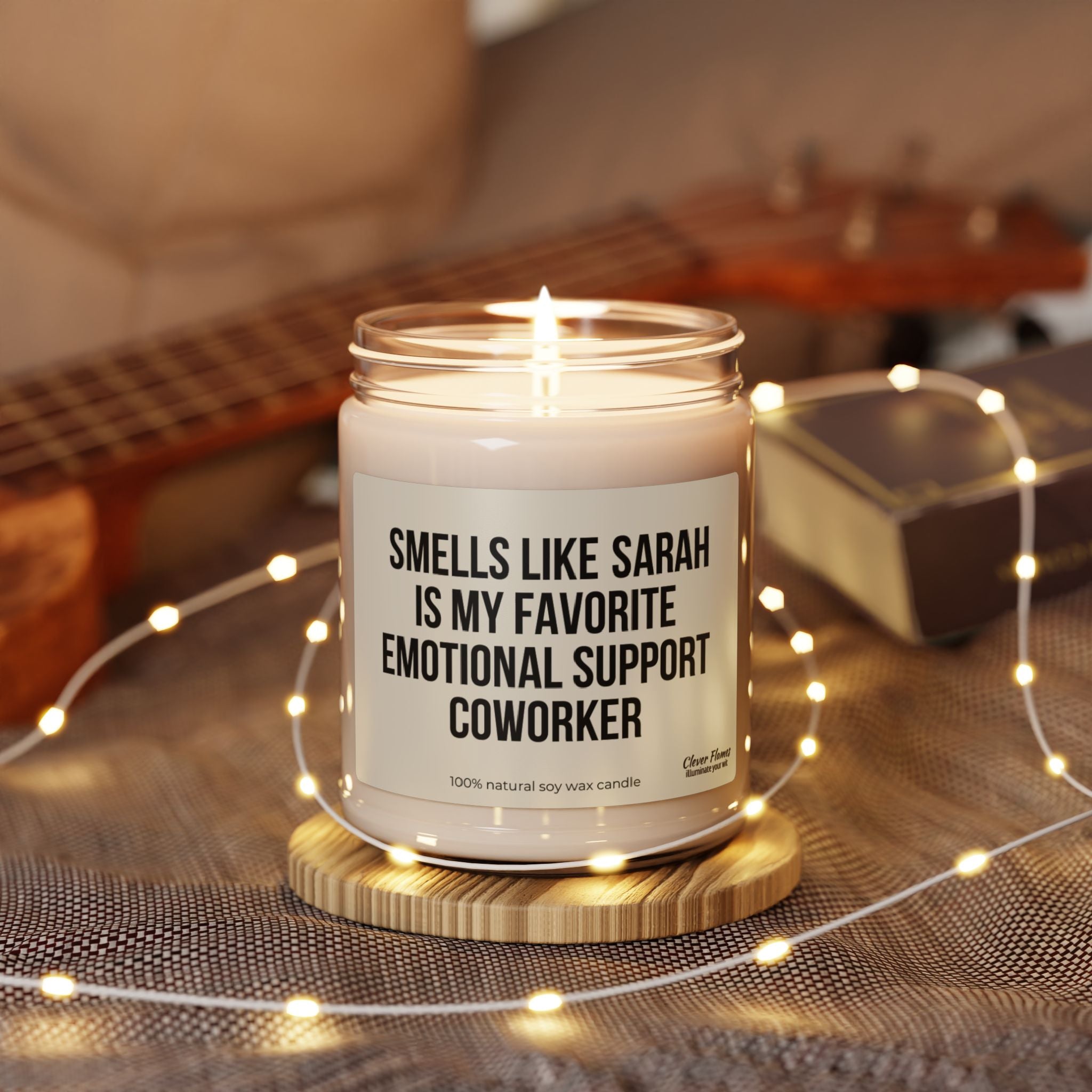 A lit candle from Printify, labeled as the "Personalized Funny Coworker Leaving Candle for Work Bestie Gift," sits on a wooden coaster. In the background, there are fairy lights, a book, and part of a ukulele, creating an eco-friendly haven.