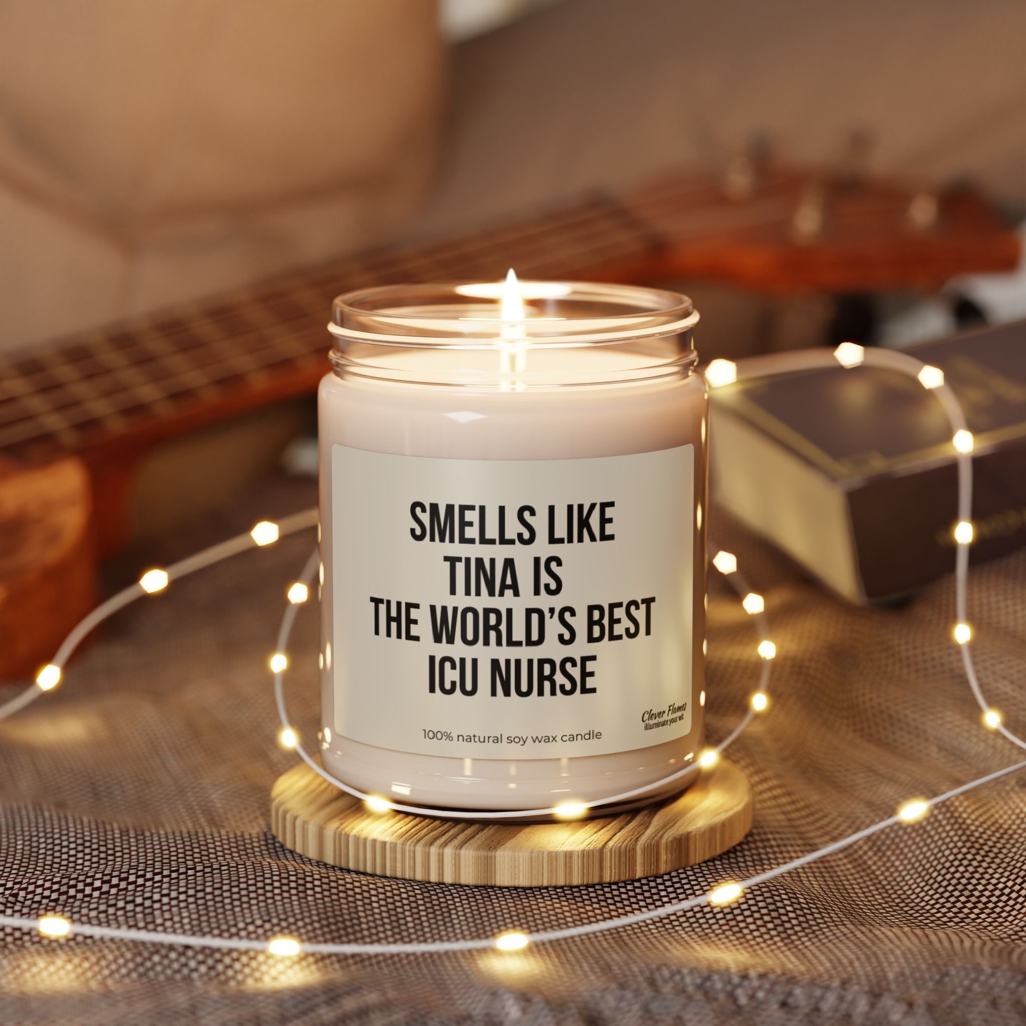 A candle from Printify, labeled "Custom Smells Like World's Best ICU Nurse," rests on a wooden coaster, making it an ideal gift for nurses. It is surrounded by fairy lights and set on a gray cloth, with a blurred ukulele and book in the background.