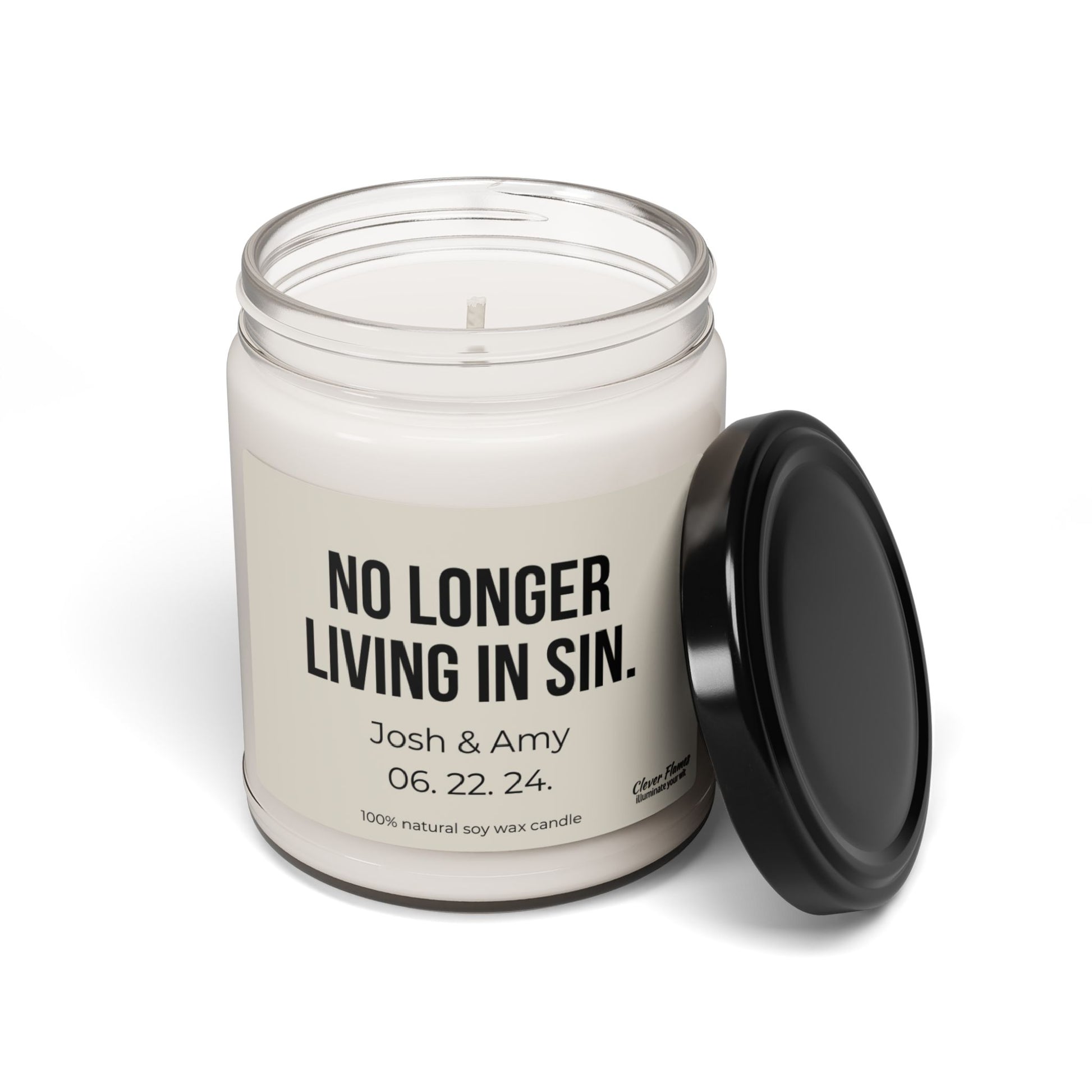 A white soy candle in a glass jar with the lid set aside displays the label "No Longer Living In Sin. Josh & Amy 06.22.24," making it an excellent personalized anniversary gift from Wedding’s product line of custom wedding gifts designed to add humor and charm for couples.