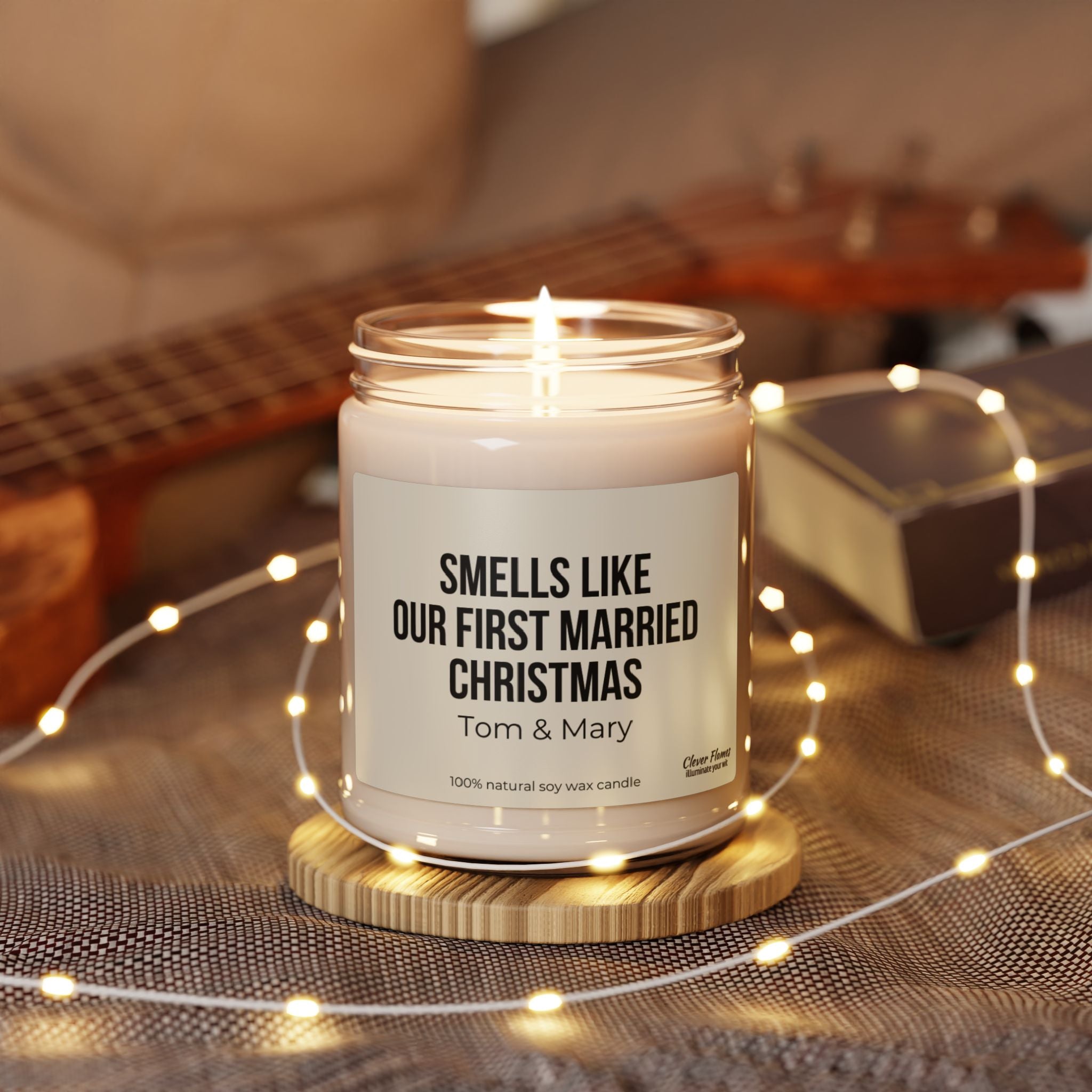 A lit Printify candle, labeled "First Married Christmas Personalized Candle Engagement Gift for Mr. and Mrs. Newlywed," is nestled in a jar adorned with the phrase "Smells Like Just Married." It rests on a wooden base surrounded by string lights, creating an ideal gift for newlyweds, with a ukulele and book softly blurred in the background.