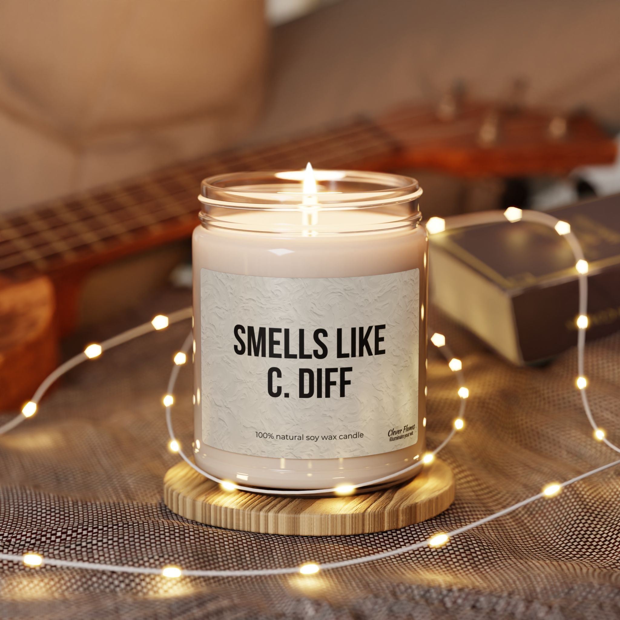 The Printify Smells Like C. Diff soy wax candle is lit on a wooden coaster, surrounded by soft string lights and a blurred background of a ukulele and box—an ideal humorous ambiance gift for nurses and medical professionals.