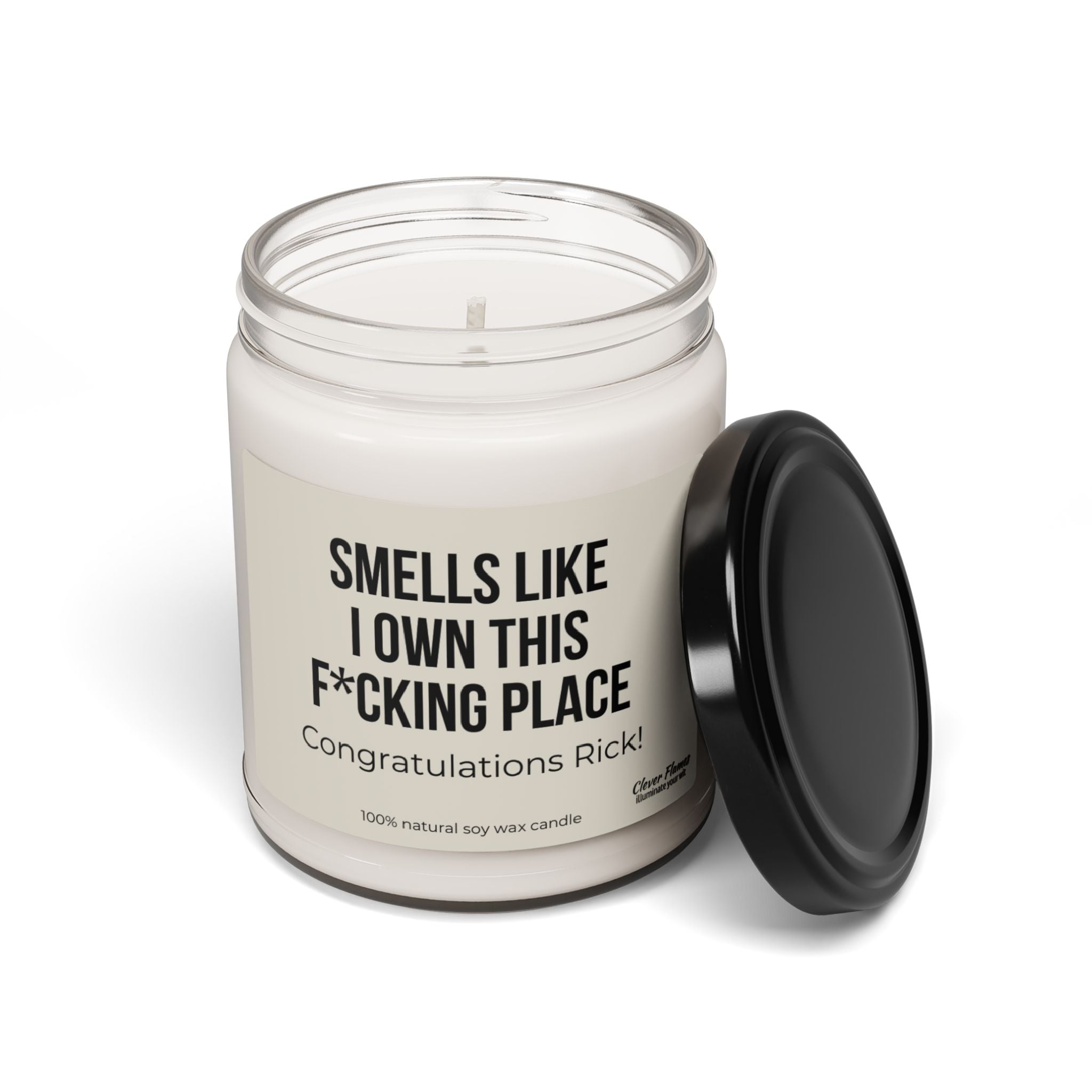 A white scented soy candle from Printify rests in a glass jar with a black lid beside it. The label reads, "SMELLS LIKE I OWN THIS F*CKING PLACE." Congratulations Rick! This candle, made from 100% natural soy wax, serves as the perfect housewarming gift.