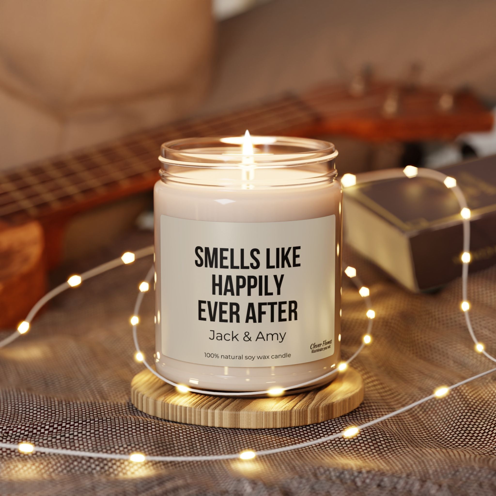 A Personalized Wedding Gift candle from the brand Wedding, labeled "Smells Like Happily Ever After," is lit and placed on a wooden coaster, surrounded by a string of warm fairy lights. In the background, a blurred wooden ukulele and a closed box complete this perfect gift for newlyweds to evoke cherished memories.