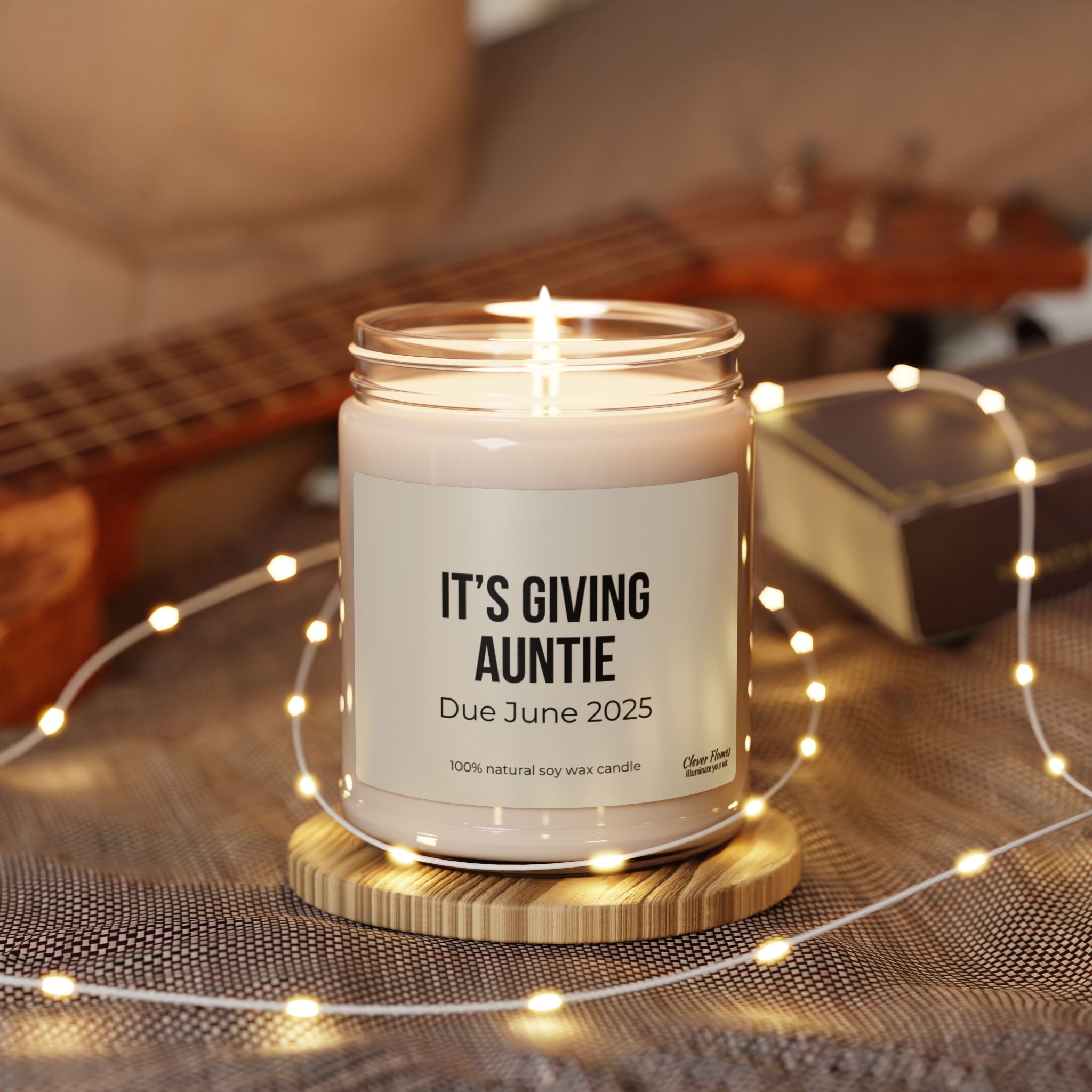 A lit Printify Pregnancy Announcement Auntie Personalized Pregnancy Reveal Candle, custom-labeled with "It's Giving Auntie, Due June 2025," rests on a wooden coaster. Crafted from 100% natural soy wax, it envelops the cozy atmosphere with warmth while a ukulele and string lights twinkle in the background—perfect for baby reveal celebrations.