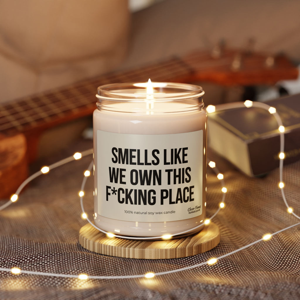 A lit Smells Like We Own This F*Cking Place Candle in a glass jar, resting on a wooden coaster surrounded by string lights.