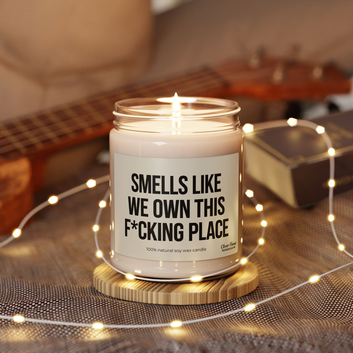 A lit Smells Like We Own This F*Cking Place Candle in a glass jar, resting on a wooden coaster surrounded by string lights.