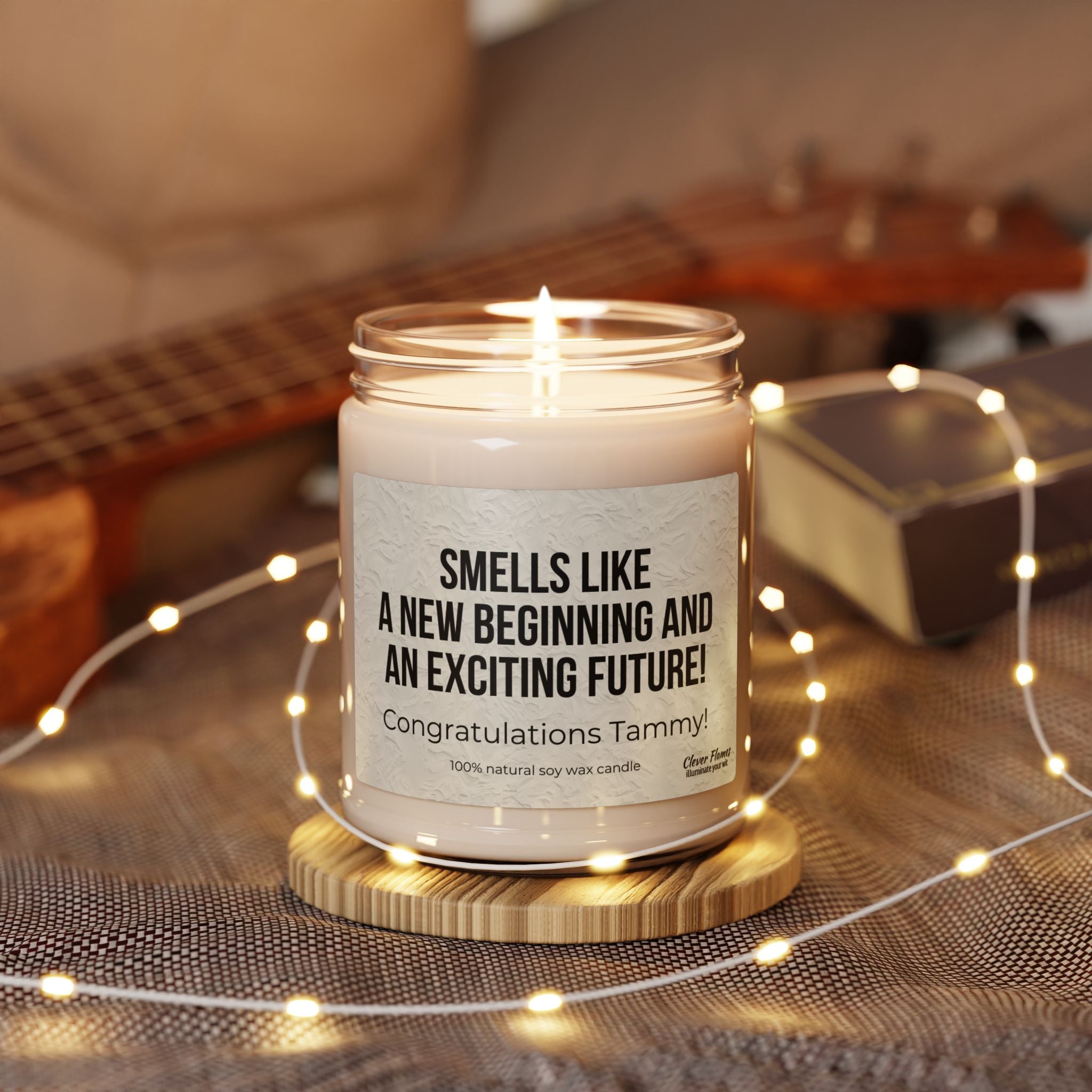 A personalized Printify candle labeled “Smells Like a New Beginning and an Exciting Future” made of natural soy wax sits on a wooden surface with string lights, while a blurred ukulele enhances this new beginnings gift. .