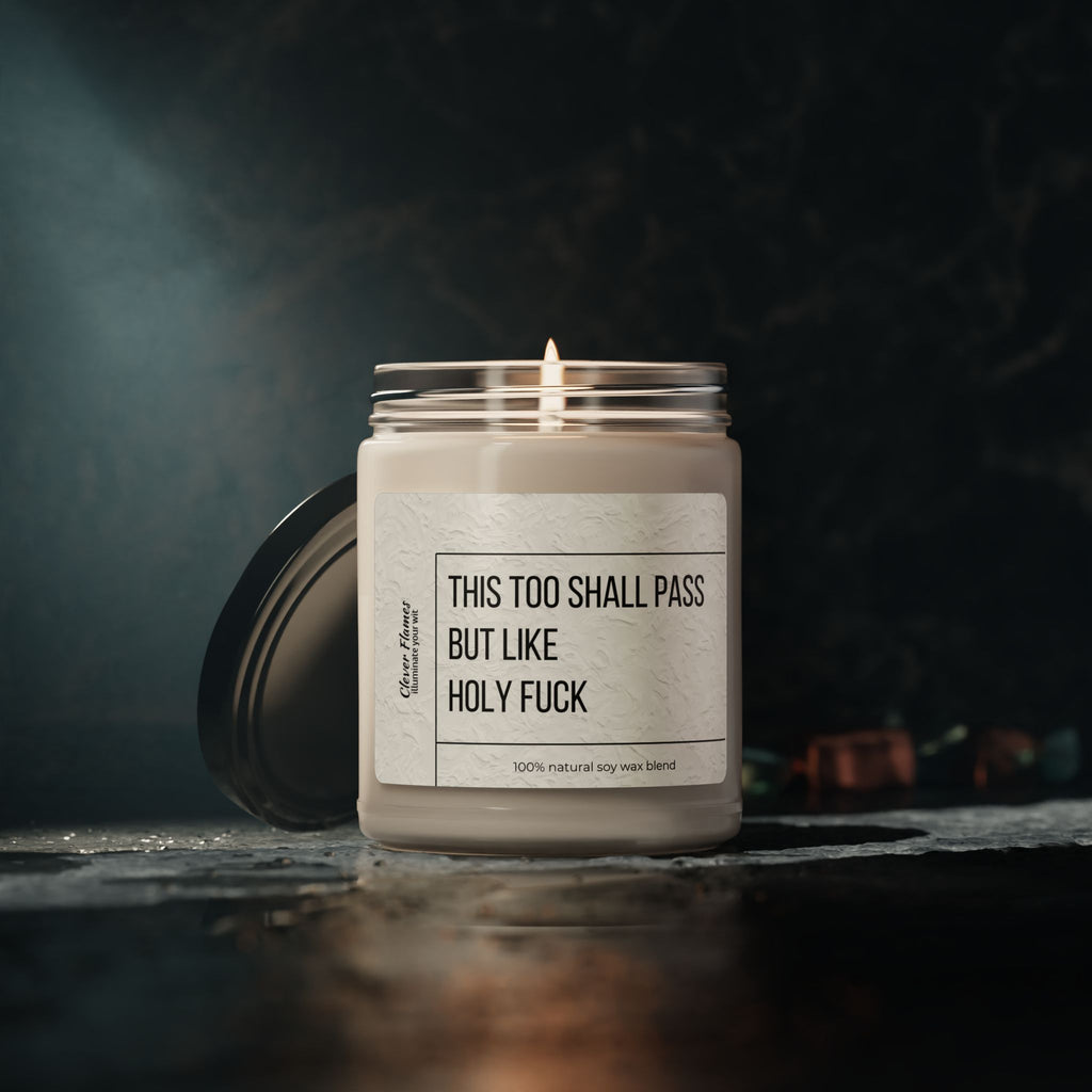 The 9oz Humorous Scented Soy Candle from Printify, labeled This Too Shall Pass, But Like Holy F***, sits in a glass jar with the lid beside it on a dark textured surface against a moody backdrop.