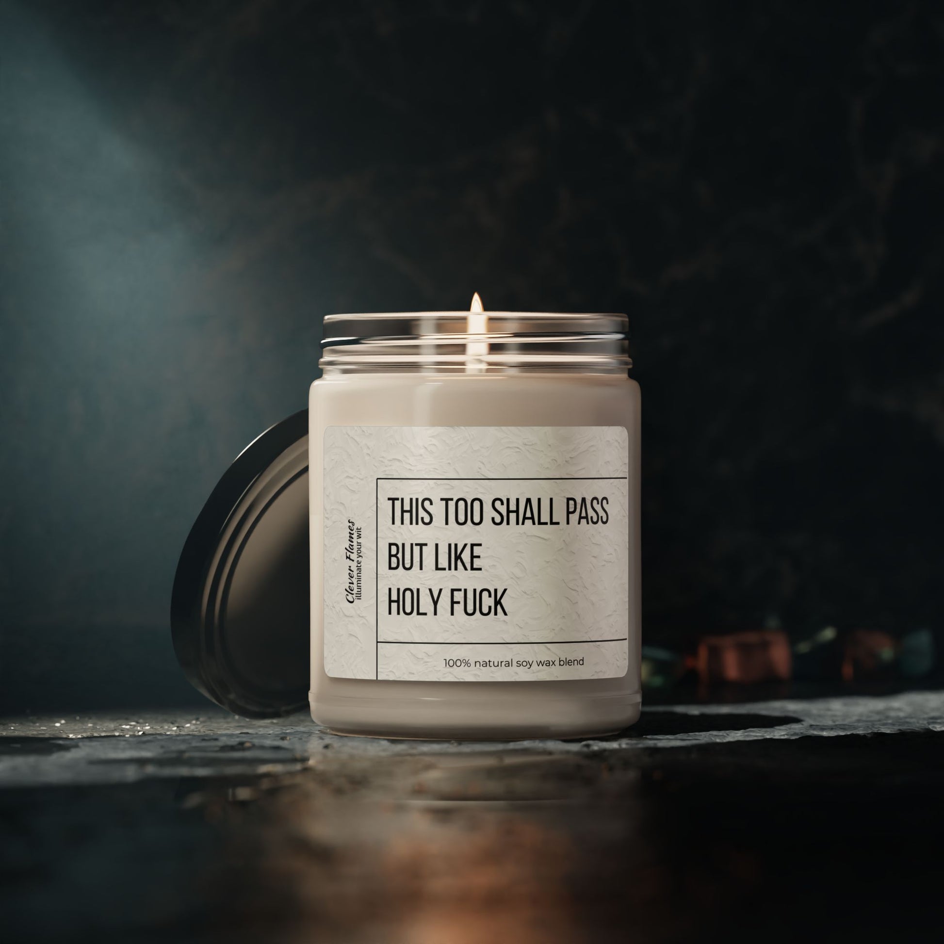 The 9oz Humorous Scented Soy Candle from Printify, labeled This Too Shall Pass, But Like Holy F***, sits in a glass jar with the lid beside it on a dark textured surface against a moody backdrop.
