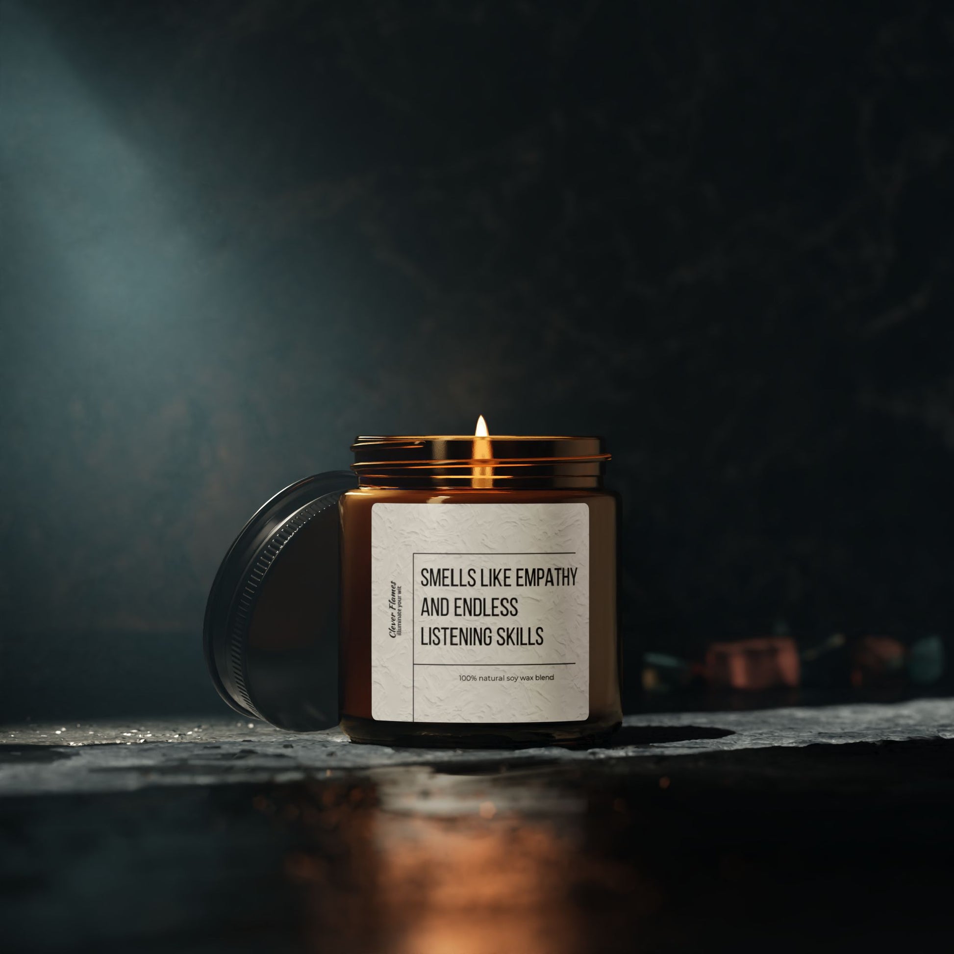 A lit, eco-friendly Counselor Candle by Printify sits on a stone surface, housed in a dark brown jar with a label that reads "Smells Like Empathy & Endless Listening Skills." The lid is removed and placed beside the jar, releasing its calming scent into the dark, moody background.