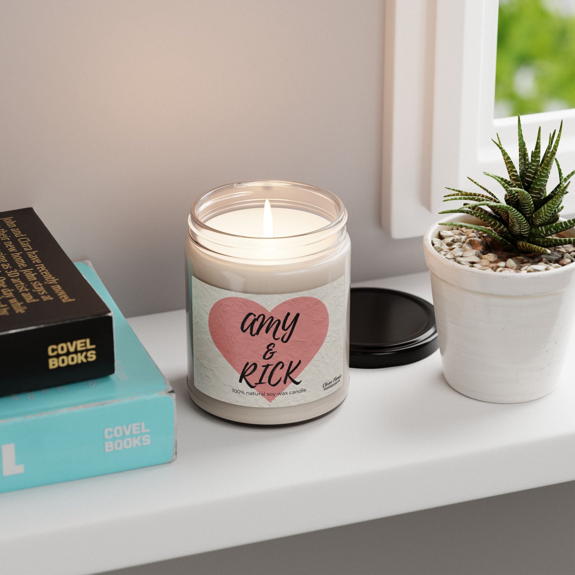 A Printify Personalized Couples Candle with Amy & Rick in a romantic heart design made from soy wax graces a white shelf near a potted succulent, beside stacked books for an ideal romantic gift setting against a backdrop of wall and window edge.