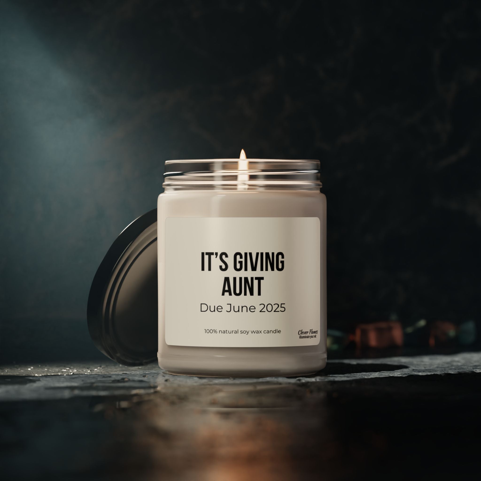 A lit candle from the Printify collection, labeled with "ITS GIVING AUNT" and "Due June 2025," rests on a dark surface. This 9oz Custom Baby Announcement Aunt Scented Soy Candle creates a cozy and warm ambiance, making it an ideal pregnancy announcement candle. Perfect as an aunt-to-be gift, it promises a natural soy wax experience.