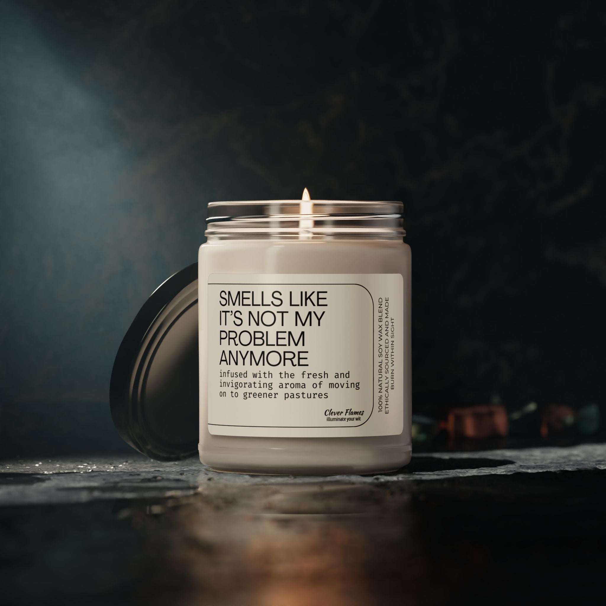 A lit candle in a jar sits on a dark surface. The label reads Smells Like Its Not My Problem Anymore, and mentions an aroma associated with moving on to greener pastures. The background is dimly lit, adding a moody ambiance.