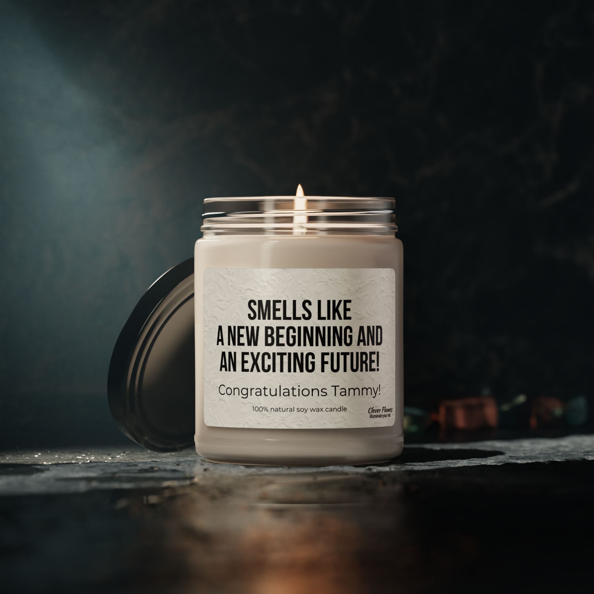 A lit Printify candle with a dark background features a label reading, Smells like a new beginning and an exciting future! Congratulations Tammy! Made of 100% natural soy wax, its lid is open beside the jar—perfect for celebrating new jobs, graduations, or fresh starts.