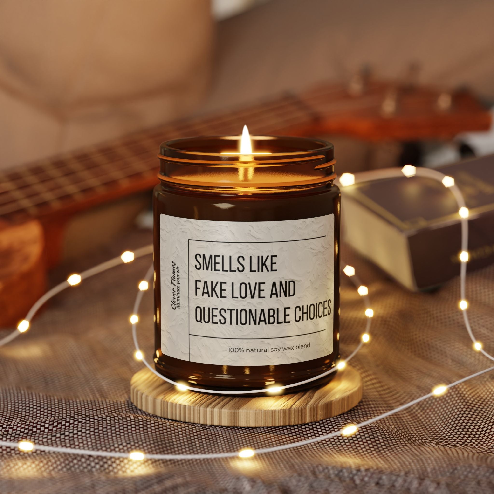 A scented soy candle in a brown jar on a wooden coaster, with the label "Smells Like Fake Love and Questionable Choices" by Printify, offers an eco-friendly touch. Adorned with string lights, it's an ideal gift for romantic occasions. In the background, a blurred guitar and closed book add to the cozy atmosphere.