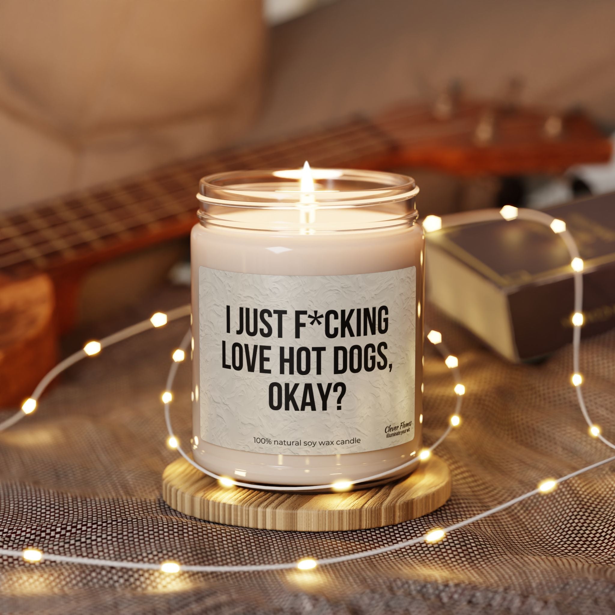 The Printify “I Just F*cking Love Hot Dogs Okay?” Scented Soy Candle (9oz) burns in a reusable glass jar on a wooden coaster, with its bold label in focus. Soft fairy lights, a guitar, and a book blur gently in the background.