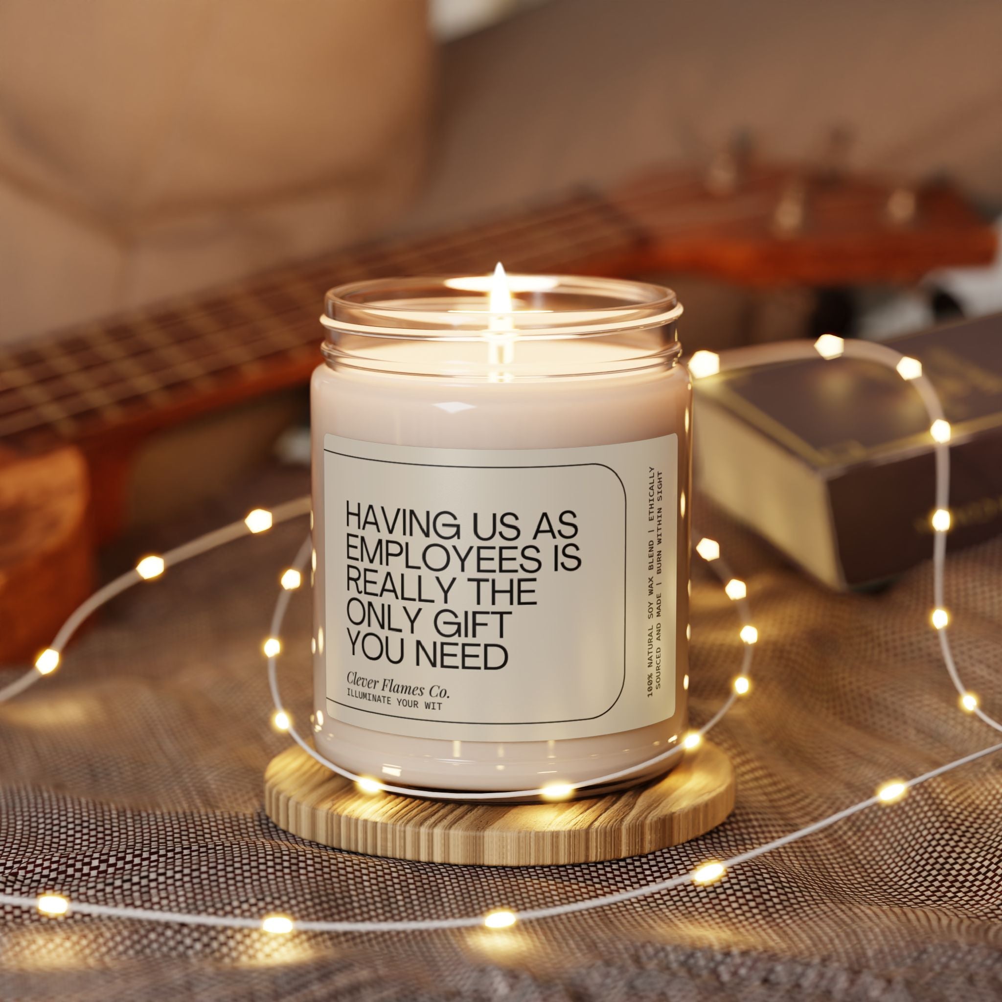 A lit Printify scented soy candle with a cinnamon vanilla scent in a glass jar is placed on a wooden coaster, encircled by string lights. The label humorously states, "Having us as employees is really the only gift you need." In the background, a guitar and a book are blurred, making it an ideal gift for boss appreciation. This is the 9oz Funny Boss Gift Scented Soy Candle, perfect for office parties or celebrating the best boss ever on Boss Day.
