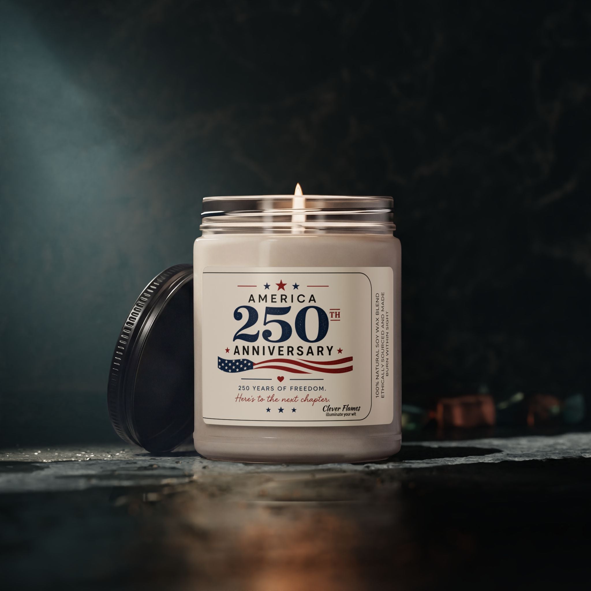 A lit Printify America 250th Anniversary Soy Candle, 9oz | Patriotic Independence Day, sits in a glass jar with its lid beside it on a dark surface. The label celebrates 250 Years of Freedom.