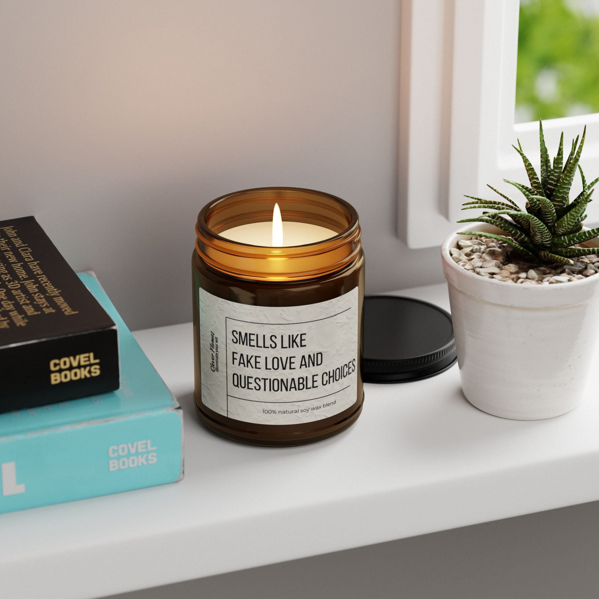 A lit brown jar candle, "Smells Like Fake Love and Questionable Choices," by Printify, ideal for Valentine's Day or relationship gifts, rests on a white shelf beside a small potted succulent and a stack of books. The scented soy candle enjoys the gentle embrace of soft daylight streaming through the nearby window.