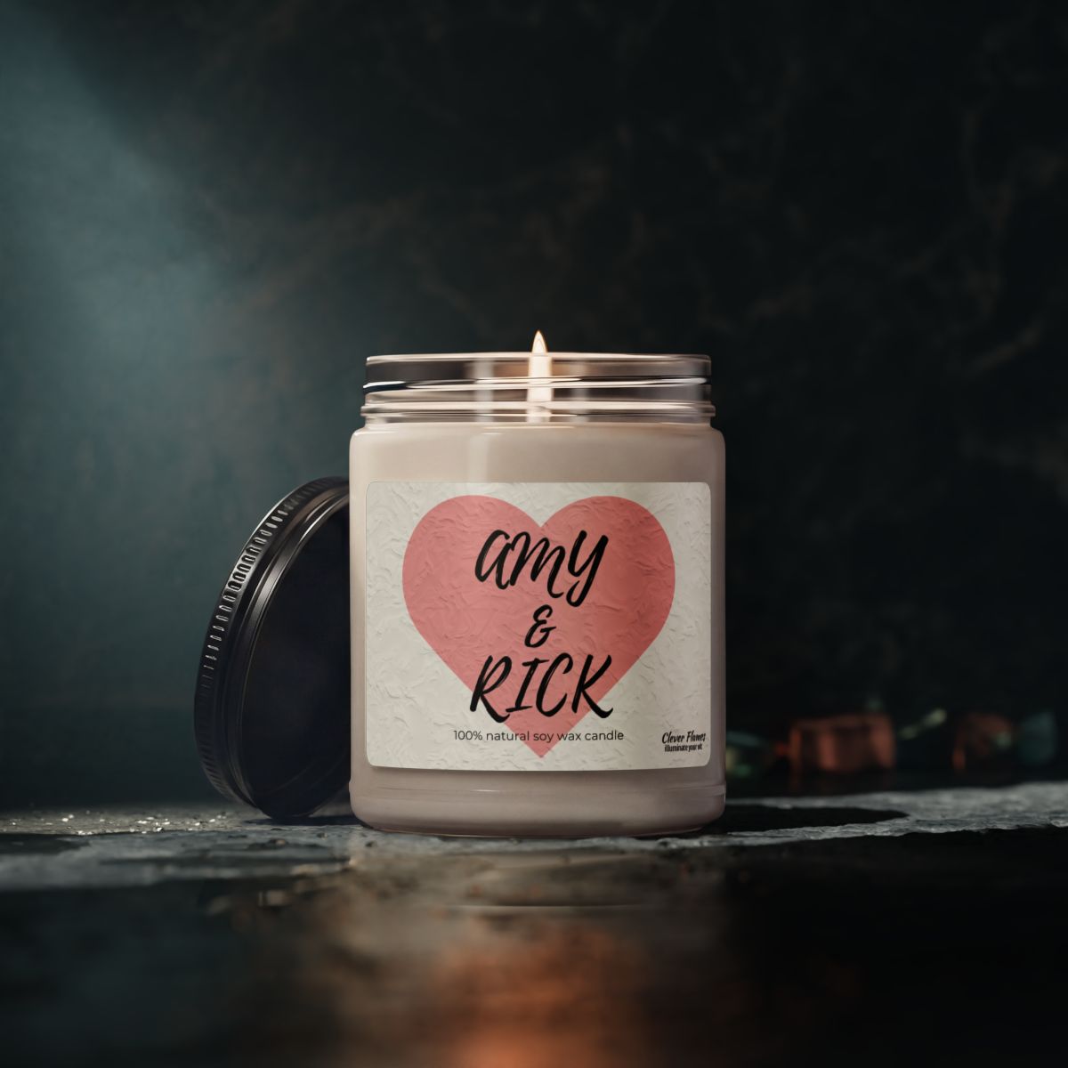 A glowing white soy candle from Printify’s 9oz Personalized Couples Candle – Romantic Heart Design collection sits in a jar adorned with a pink heart label that reads “Amy & Rick,” making it an ideal custom gift for couples. The jar’s black lid rests nearby on a dark, textured surface.