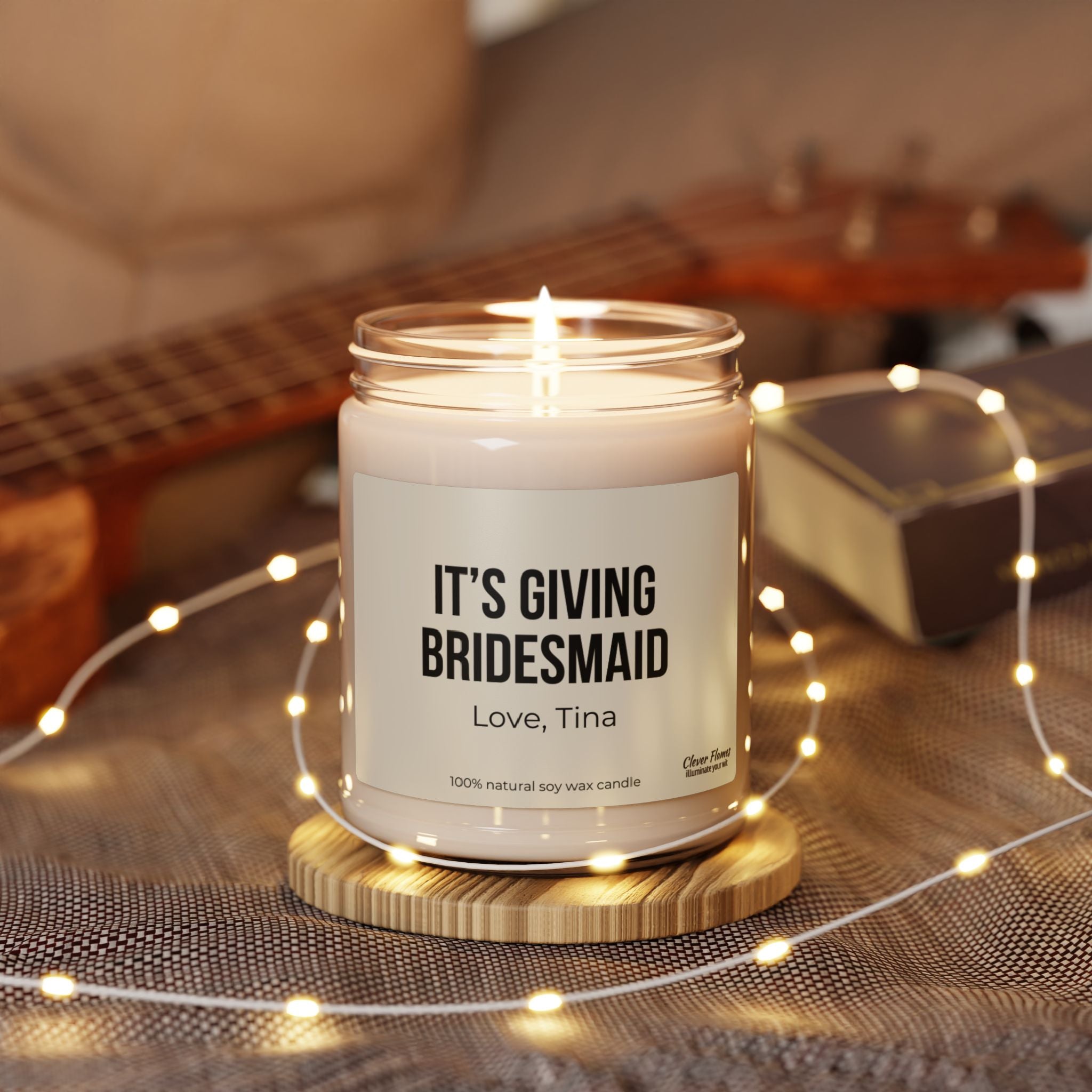 A personalized soy wax candle, branded as Wedding's "It's Giving Bridesmaid" gift, sits elegantly on a wooden stand. Warm white fairy lights are scattered around, and a ukulele is partially visible in the background, making it an ideal bridesmaid gift that creates a cozy atmosphere.