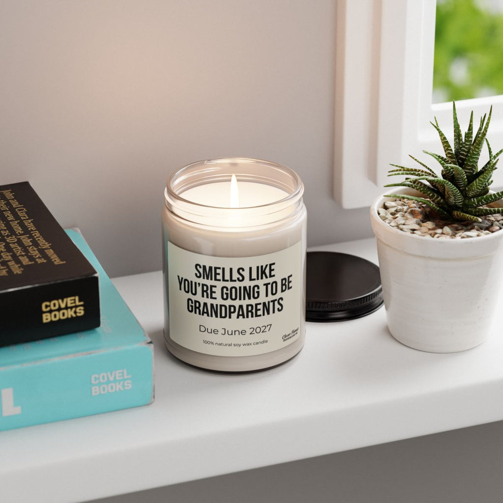A lit Printify “You’re Going to Be Grandparents” Personalized Pregnancy Announcement Candle (9oz) rests on a windowsill, its label reading: Smells like you’re going to be grandparents. Due June 2027. Nearby, a small potted plant and two books add a cozy touch—making this candle the perfect grandparent gift or unique personalized baby reveal for your loved ones.