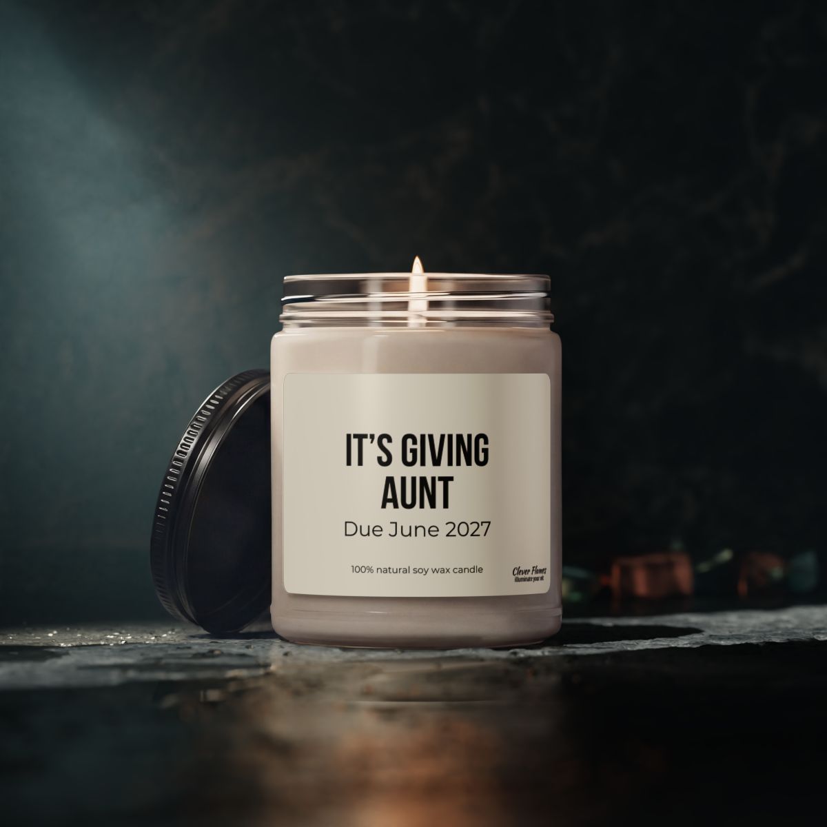 The Personalized Aunt Baby Announcement Candle – Pregnancy Reveal Gift for Aunt to Be by Printify is shown lit in a glass jar with a label that reads, “ITS GIVING AUNT Due June 2027.” Crafted from 100% natural soy wax, this 9oz candle features the lid off and resting beside the jar on a dark surface, making it the perfect personalized pregnancy announcement or baby reveal gift for your special aunt-to-be.