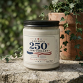 The America 250th Anniversary Soy Candle, 9oz by Printify—featuring patriotic designs—rests on a stone surface next to a terracotta pot with ivy, making it an ideal Semiquincentennial gift for any Independence Day celebration.