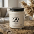 A Printify America 250th Anniversary Scented Soy Candle, 9oz, with a patriotic label reading “250 years of freedom. Here’s to the next chapter,” sits in a glass jar on a wooden table, illuminated by gentle sunlight.