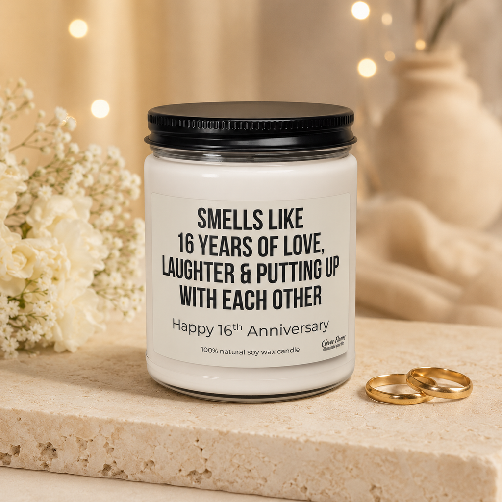 A white “16 Years of Love, Laughter & Putting Up With Each Other” candle by Printify, designed for a 16th wedding anniversary gift, rests on a stone surface beside two gold rings and white flowers in the background.