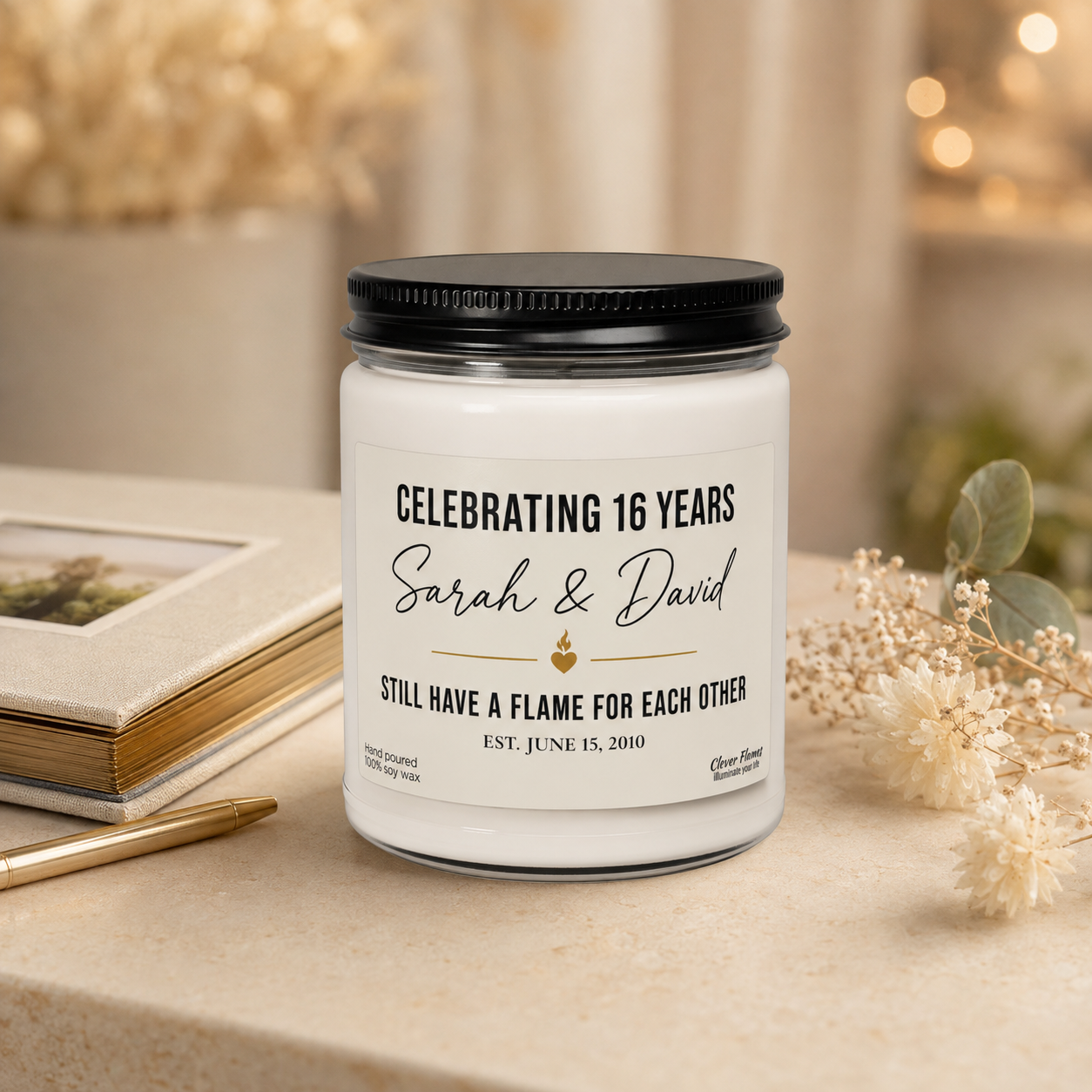 A Personalized 16th Anniversary Candle from Printify sits on the table, its custom label displaying: 16 Years Married. Sarah & David. Still have a flame for each other. Est. June 15, 2010. With dried flowers and a photo frame in the background, this custom wax anniversary gift is the perfect way to celebrate your 16th wedding anniversary with your wife or husband.