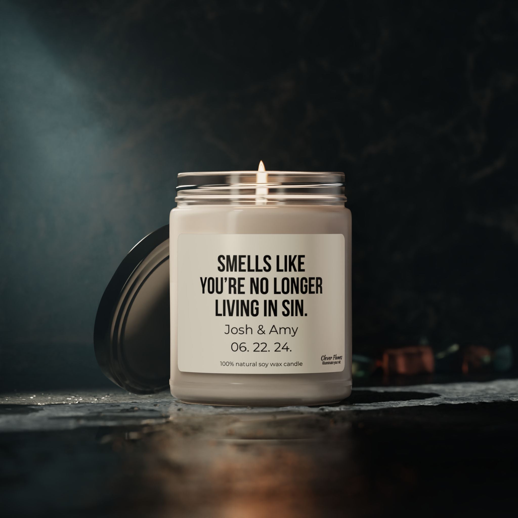 A lit soy candle from Wedding features a humorous message, "Smells like you're no longer living in sin," along with the personalization "Josh & Amy 06.22.24." Presented against a dark, moody background with its lid beside it, this candle makes an ideal engagement gift.