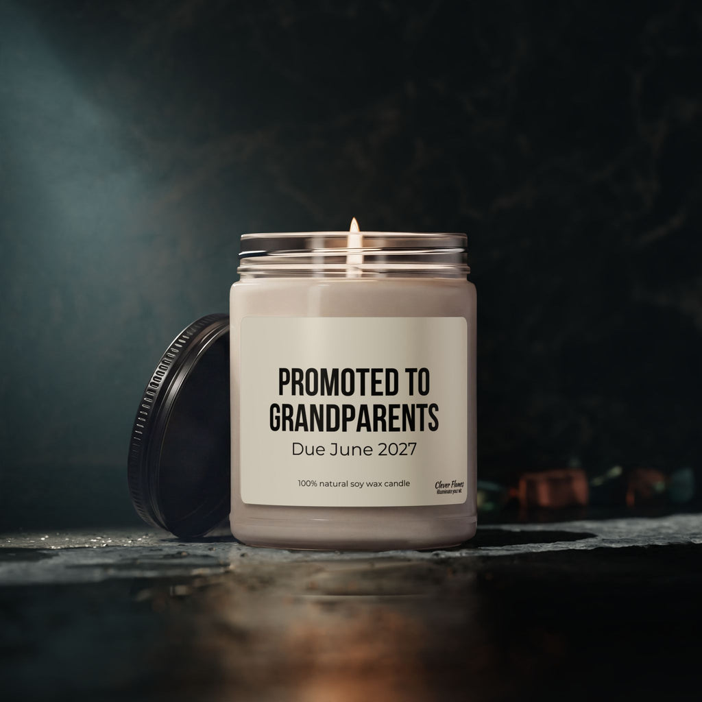 A Printify ‘Promoted to Grandparents’ 9oz scented soy candle, featuring a “Due June 2027” label, sits in its reusable glass jar with the lid resting nearby on a dark surface and a softly lit background.