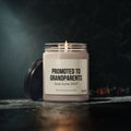 A Printify ‘Promoted to Grandparents’ 9oz scented soy candle, featuring a “Due June 2027” label, sits in its reusable glass jar with the lid resting nearby on a dark surface and a softly lit background.