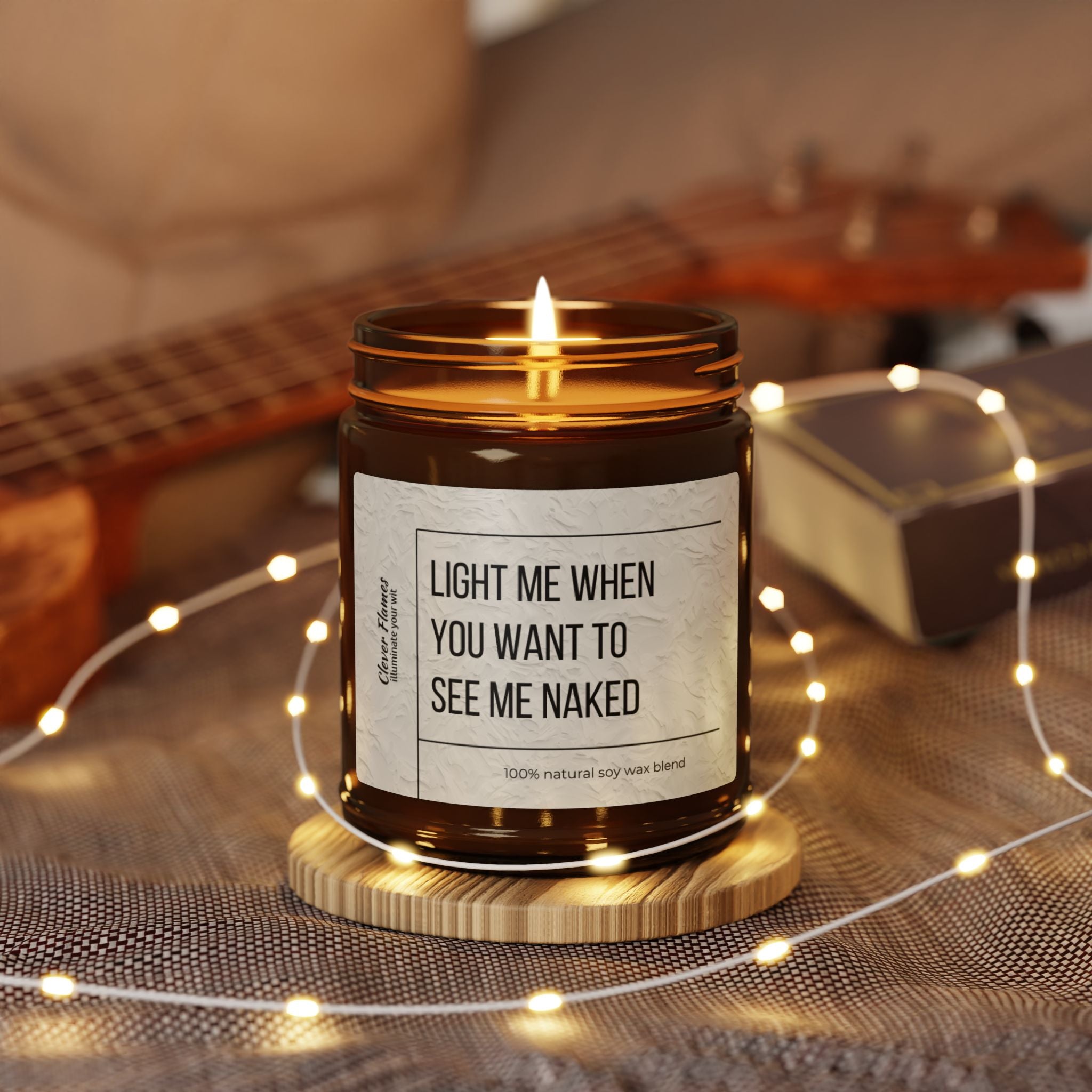 A burning "Light Me When You Want to See Me Naked" soy wax candle from Printify, housed in a brown jar with a playful label, makes for the perfect Valentine's Day gift. In the background, a ukulele and fairy lights enhance the cozy home decor atmosphere.