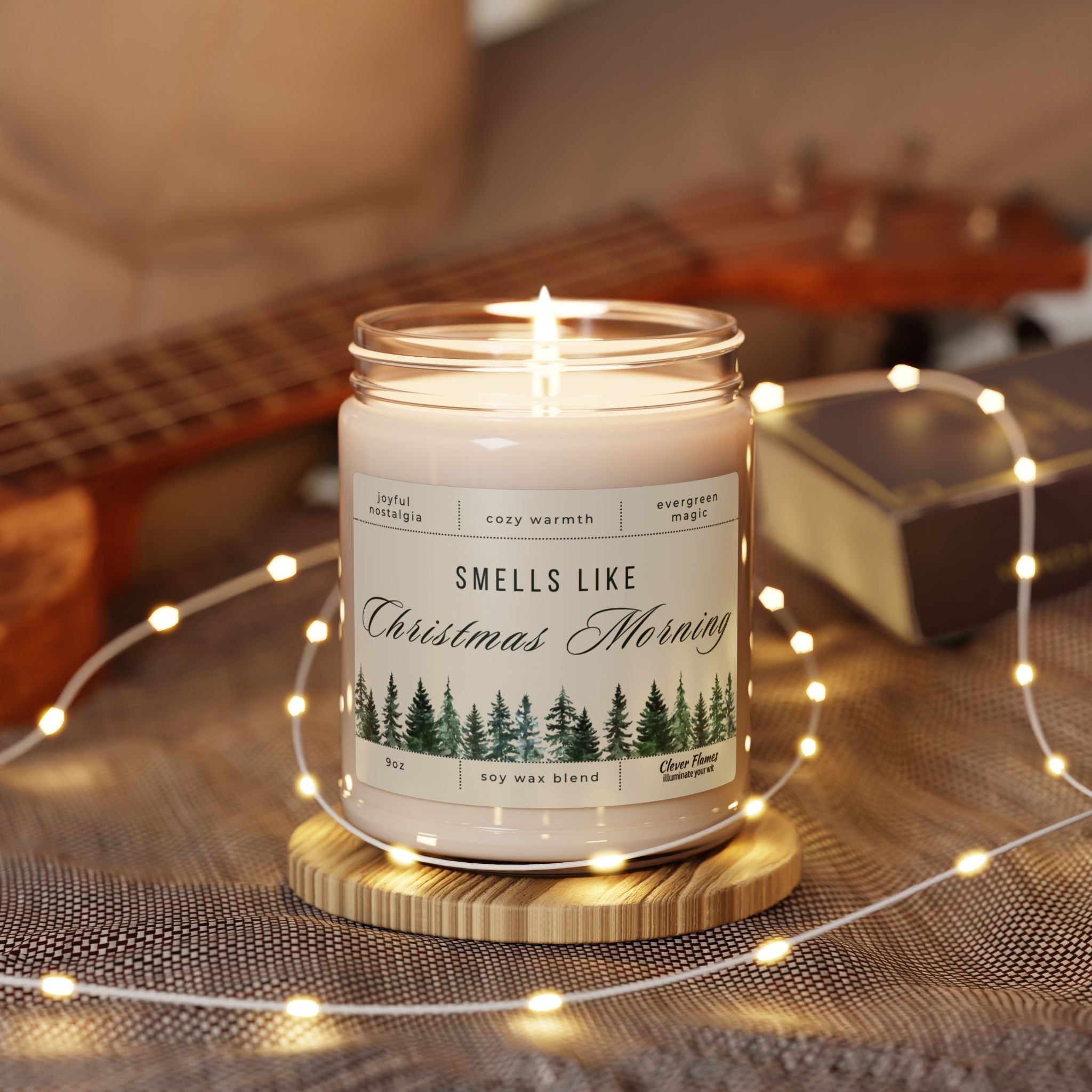 The Smells Like Christmas Morning – Holiday Soy Candle (9oz) by Printify rests on a wooden coaster among string lights, with a blurred guitar and book behind it. Its label displays pine trees and festive holiday fragrance notes.