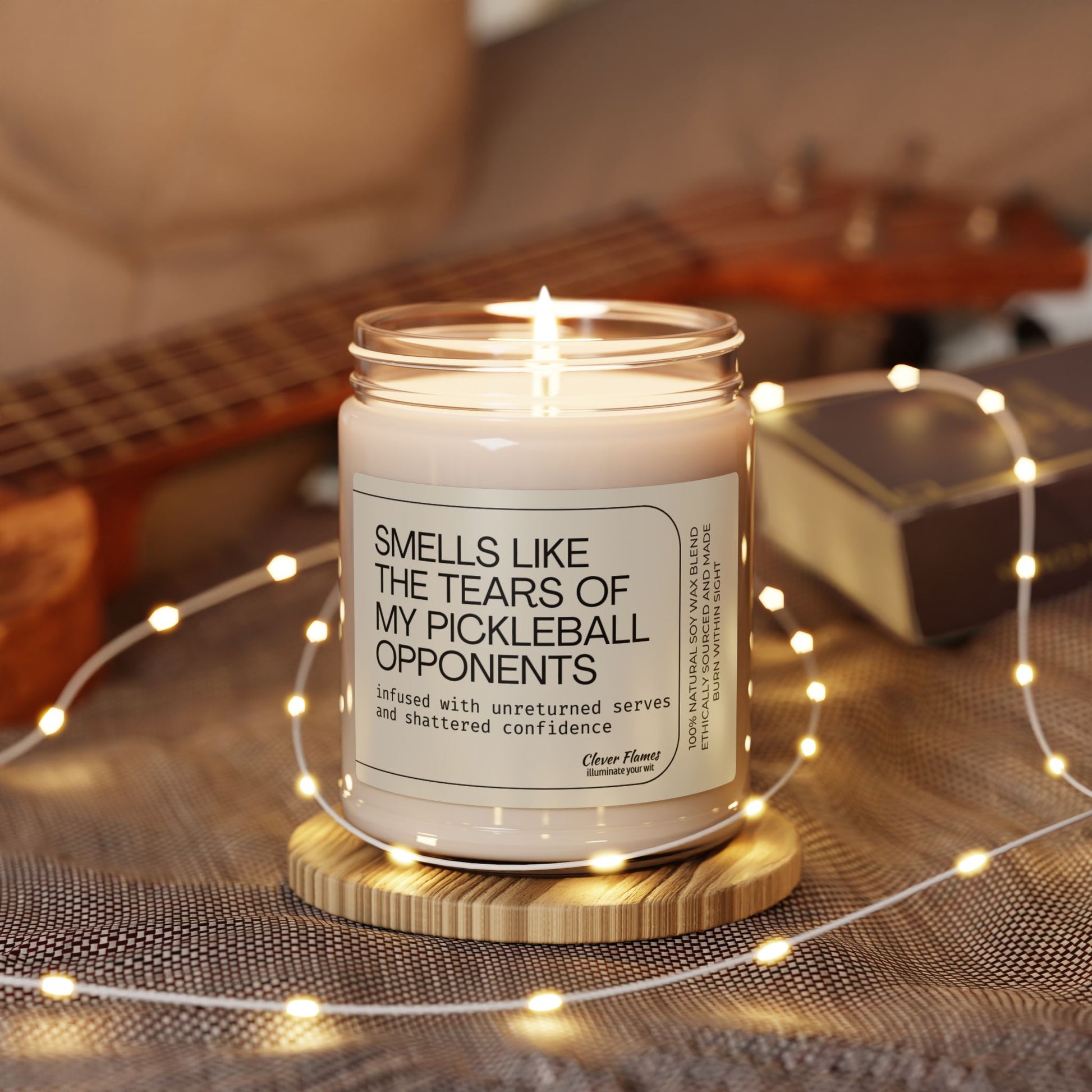 The Printify Scented Soy Candle - Smells Like the Tears of My Pickleball Opponents is an eco-friendly gift for pickleball lovers, displayed in a glass jar on a wooden coaster with cozy string lights—ideal for relaxing vibes.