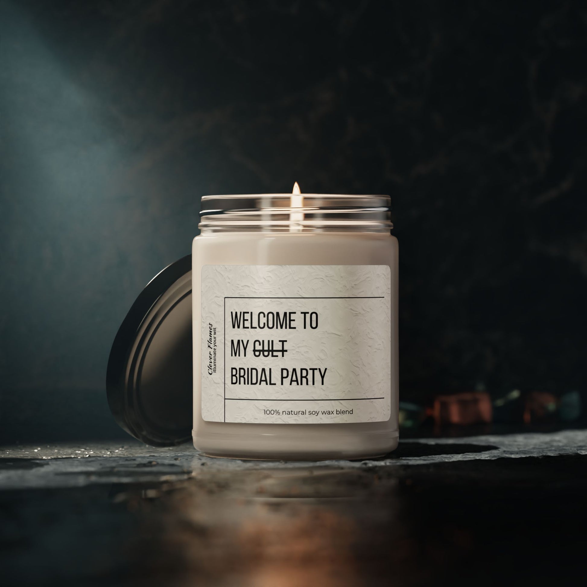 A lit Printify Welcome to My Cult Bridal Party Scented Soy Candle (9oz) sits in a glass jar with its lid beside it on a dark surface. The dim, blurred background highlights this eco-friendly candle and its unique scents.