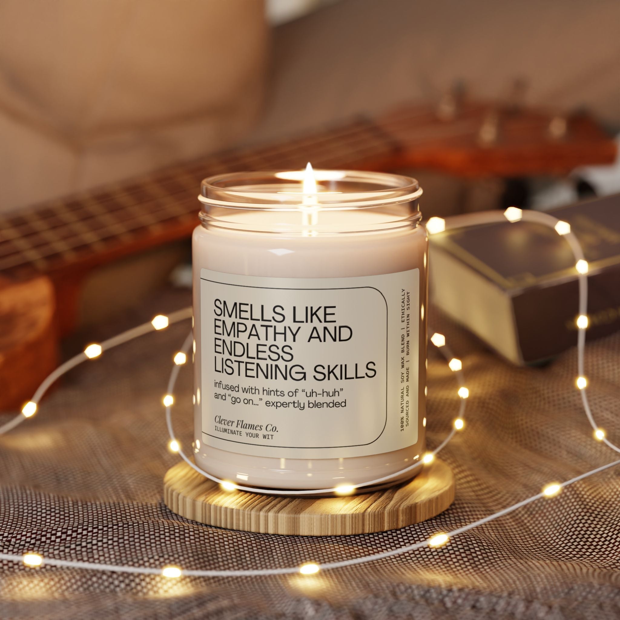 A lit soy candle from Printify, featuring a label that reads "School Counselor Candle, Guidance Counselor Gift, Thank You Gift," sits on a wooden coaster surrounded by soft fairy lights. Ideal as a Christmas gift or for funny therapist moments, this empathy-scented candle rests with a blurred guitar in the background, creating a cozy atmosphere.