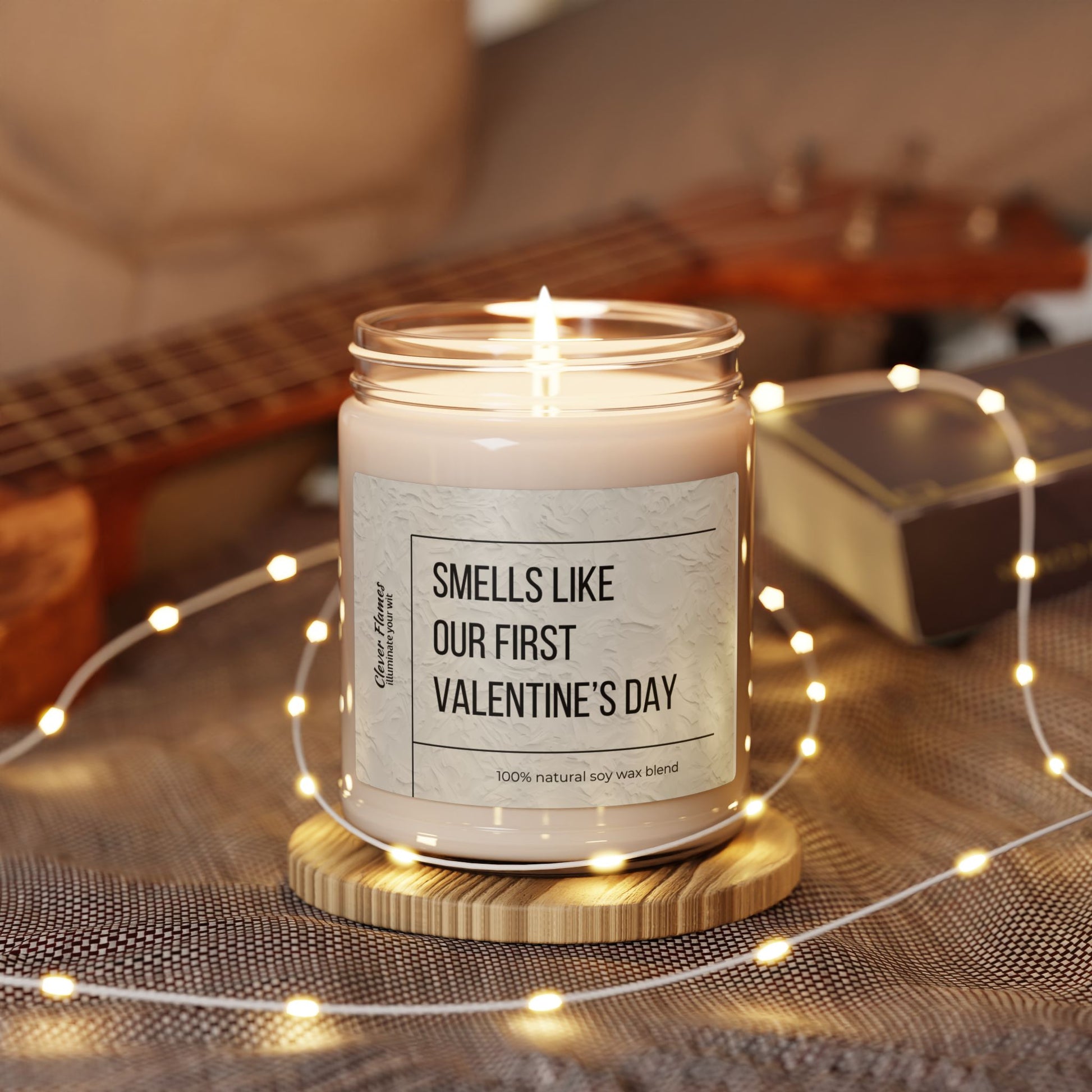 On a wooden coaster, a Printify glass jar candle labeled Smells Like Our First Valentines Day glows with natural soy wax. Its encircled by fairy lights, while a guitar and book blur in the backdrop, adding coziness.