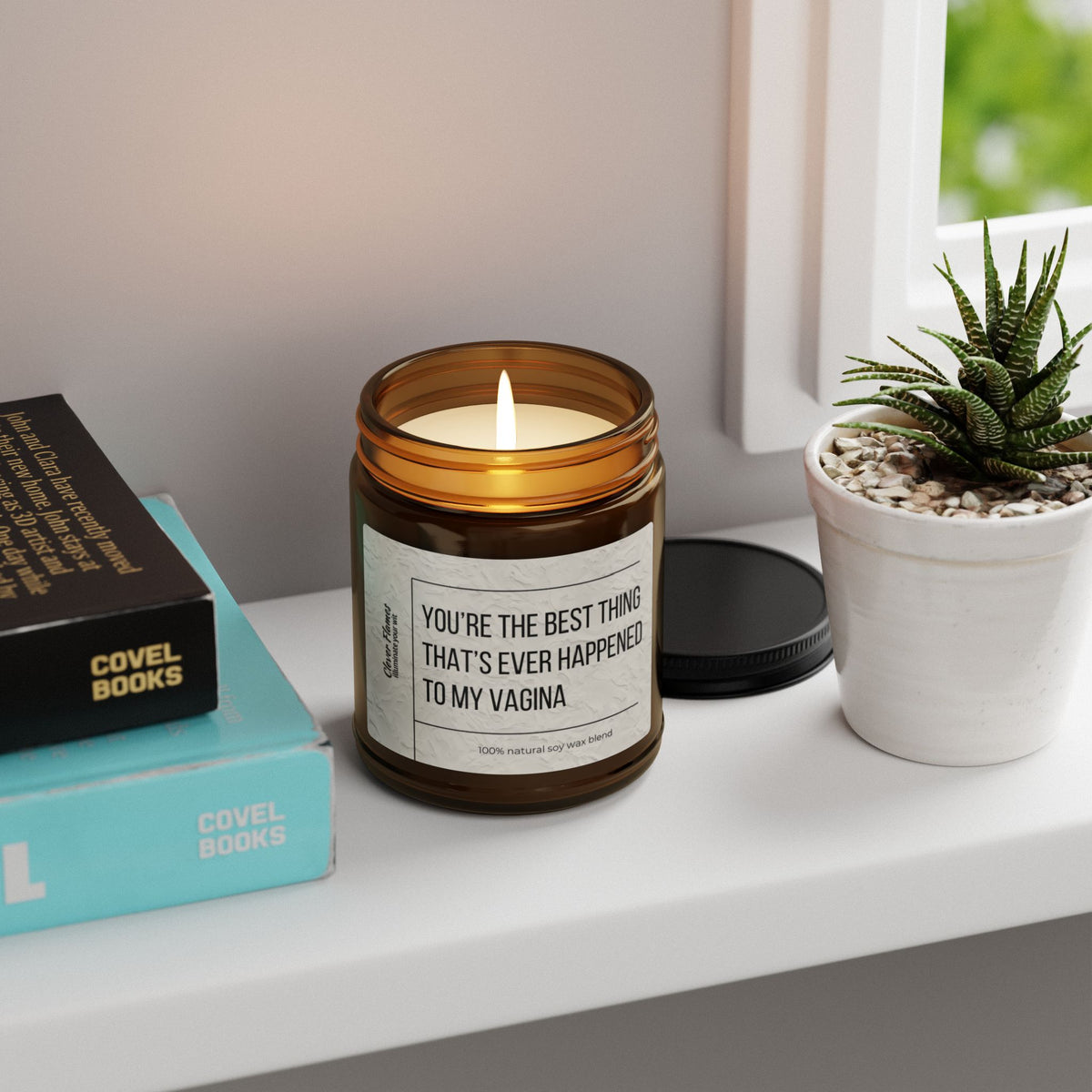 A lit brown scented soy candle with the humorous label Best Thing Thats Ever Happened To My Vagina Sex Candle Gift by Printify sits on a white shelf. Nearby, books in teal and black stack alongside a potted succulent, while the window offers a greenery view, evoking Atlantis Whispers air.