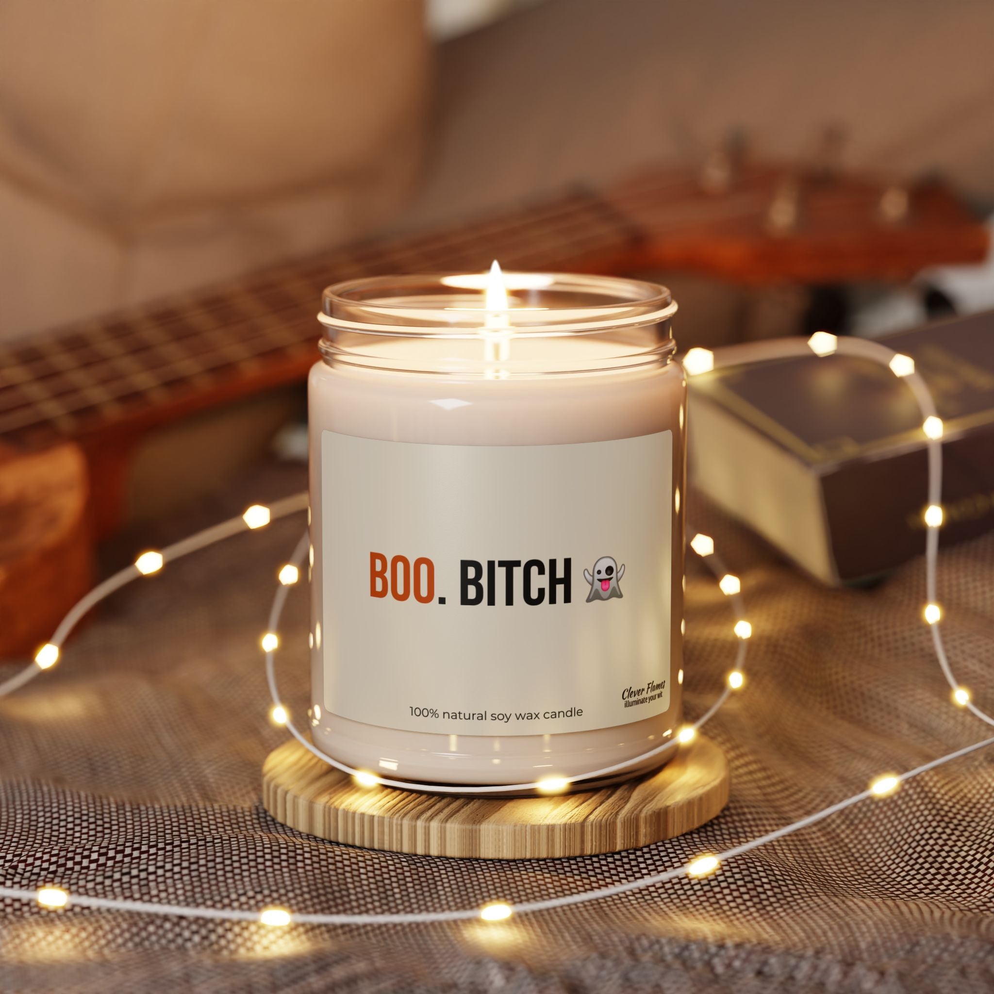A Printify Funny Halloween Fall Candle Decor labeled “Boo Bitch” with a ghost emoji rests on a wooden coaster, encircled by string lights. With a softly blurred guitar and book in the background, this spooky, sarcastic candle makes the perfect autumn gift or party favor for her or your bestie to set the mood on cozy fall nights.
