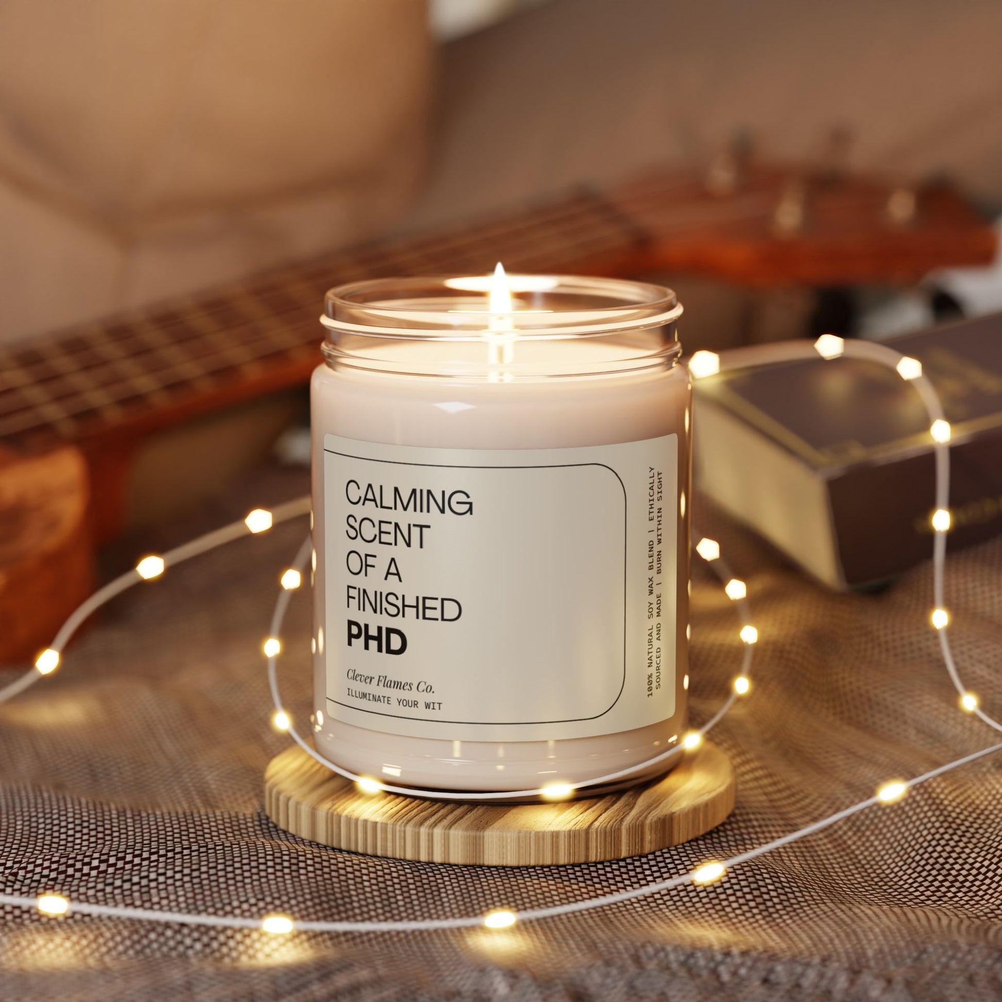 A lit Printify Finished PhD candle, humorously labeled as a graduation gift for a college graduate or master's degree recipient, sits on a wooden coaster amidst softly glowing string lights. A ukulele and a closed book blur in the background, creating a cozy atmosphere—ideal for relaxation as the perfect graduation gift.