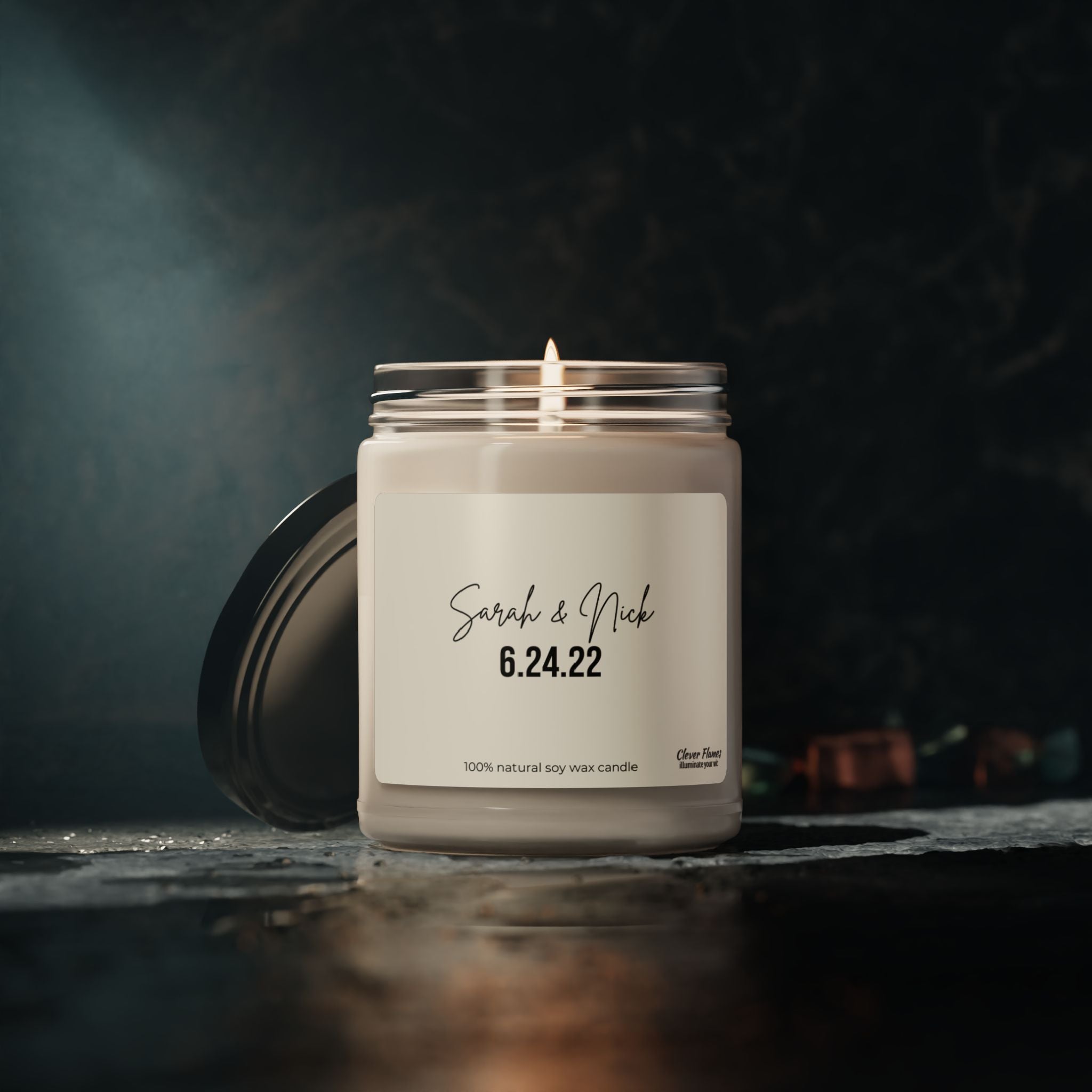 A lit candle from the Wedding brand, personalized for a wedding and placed in a glass jar, rests on a dark surface. The label displays the names Sarah & Nick along with the date 6.24.22, emphasizing its 100% natural soy wax composition. This custom candle creates a warm and inviting atmosphere with its gentle glow, ideal for cherished moments.