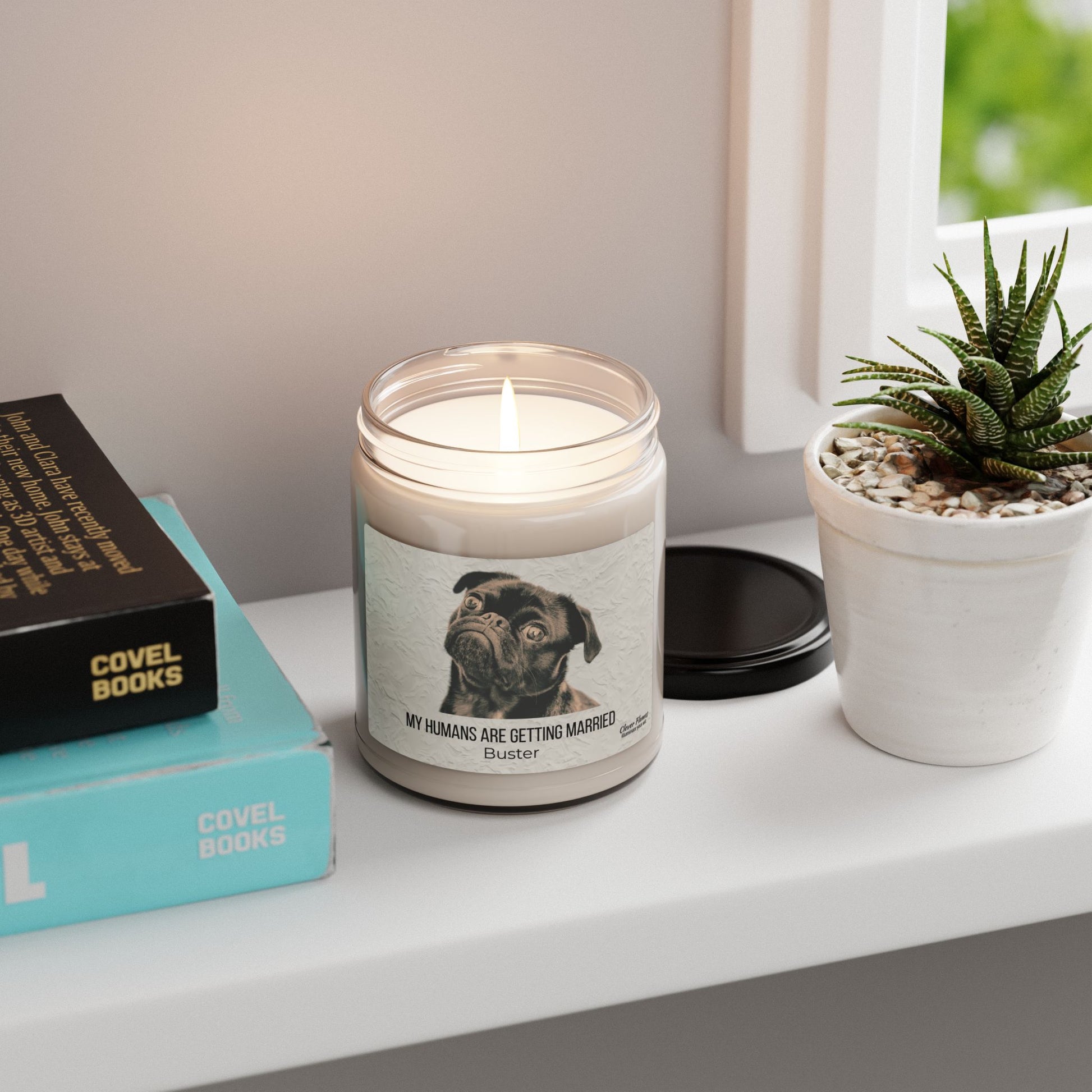 The My Humans Are Getting Married Personalized Pet Engagement Candle by Printify, featuring a pug and custom text, sits on a white shelf with a succulent and books—an adorable engagement gift for pet lovers.