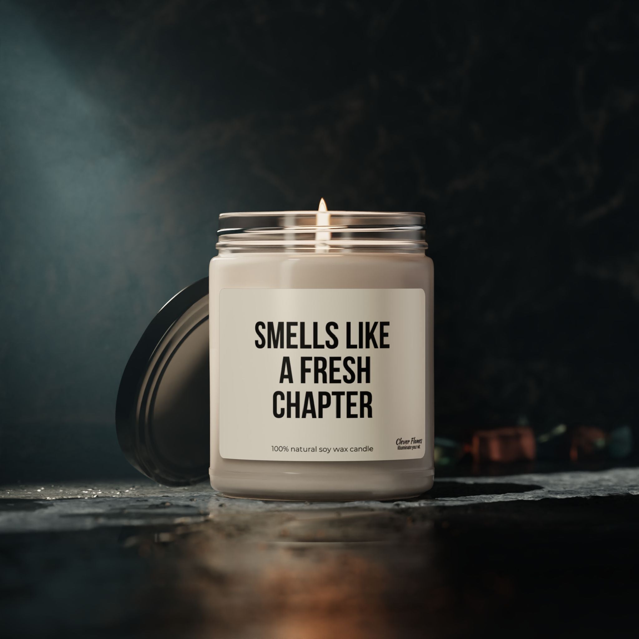 A candle from Printify's collection, labeled "Funny Candle For Reader Gift Smells Like a Fresh Chapter for Romance Reader Bookshelf Decor Bookish Merch for Booklover Bookish," sits in a glass jar on a dark, textured surface. The dimly lit background creates a cozy and inviting atmosphere, perfect for fans of bookish candles.