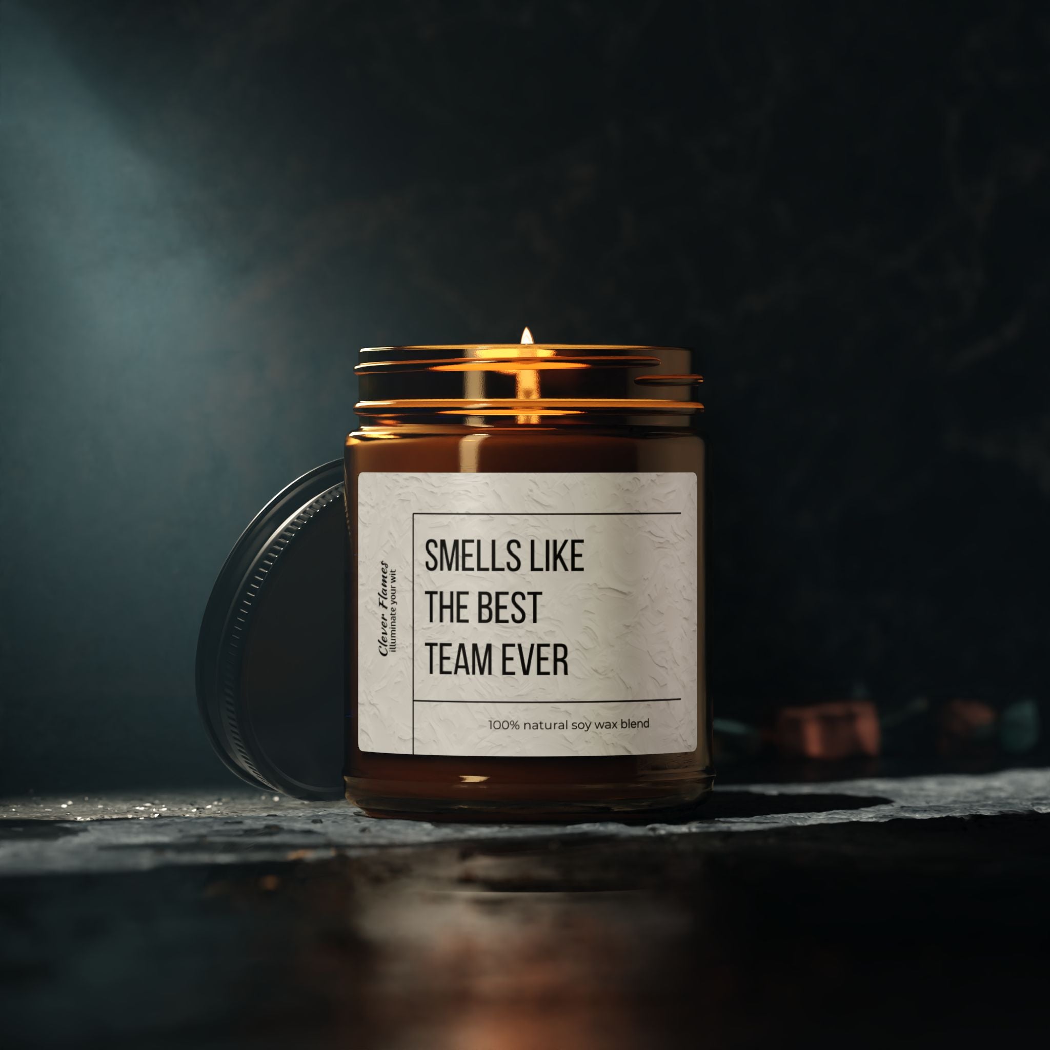 A Scented Soy Candle from Printify, resting on a dark surface in a brown glass jar, fills the air with fragrance. The label reads "BEST TEAM EVER" in bold letters, followed by "100% natural soy wax blend" in script text. Ideal for eco-friendly corporate gifts, the jar's lid leans against it in the softly lit background.