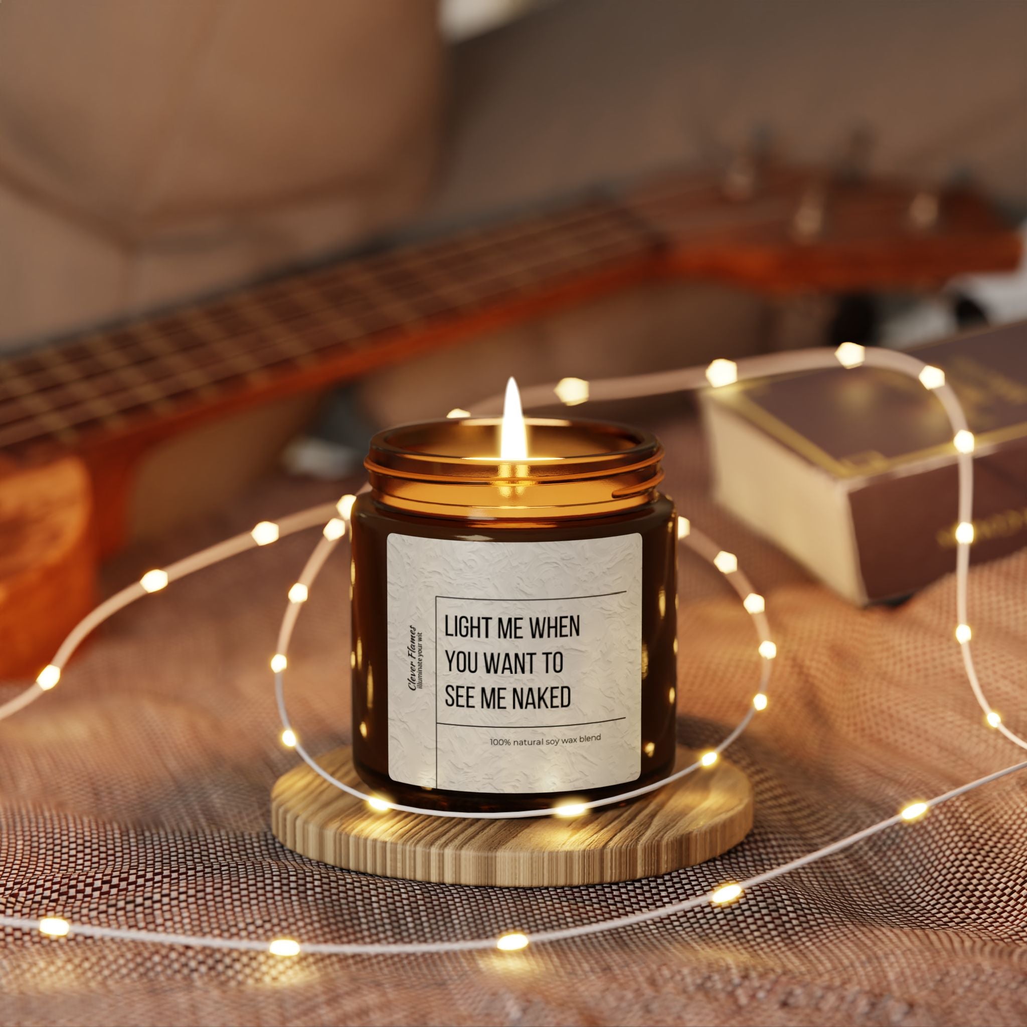 A Printify "Light Me When You Want to See Me Naked" soy wax candle, humorously labeled, rests inside a glass jar on a wooden coaster surrounded by string lights. In the background, a guitar and book create a romantic atmosphere.