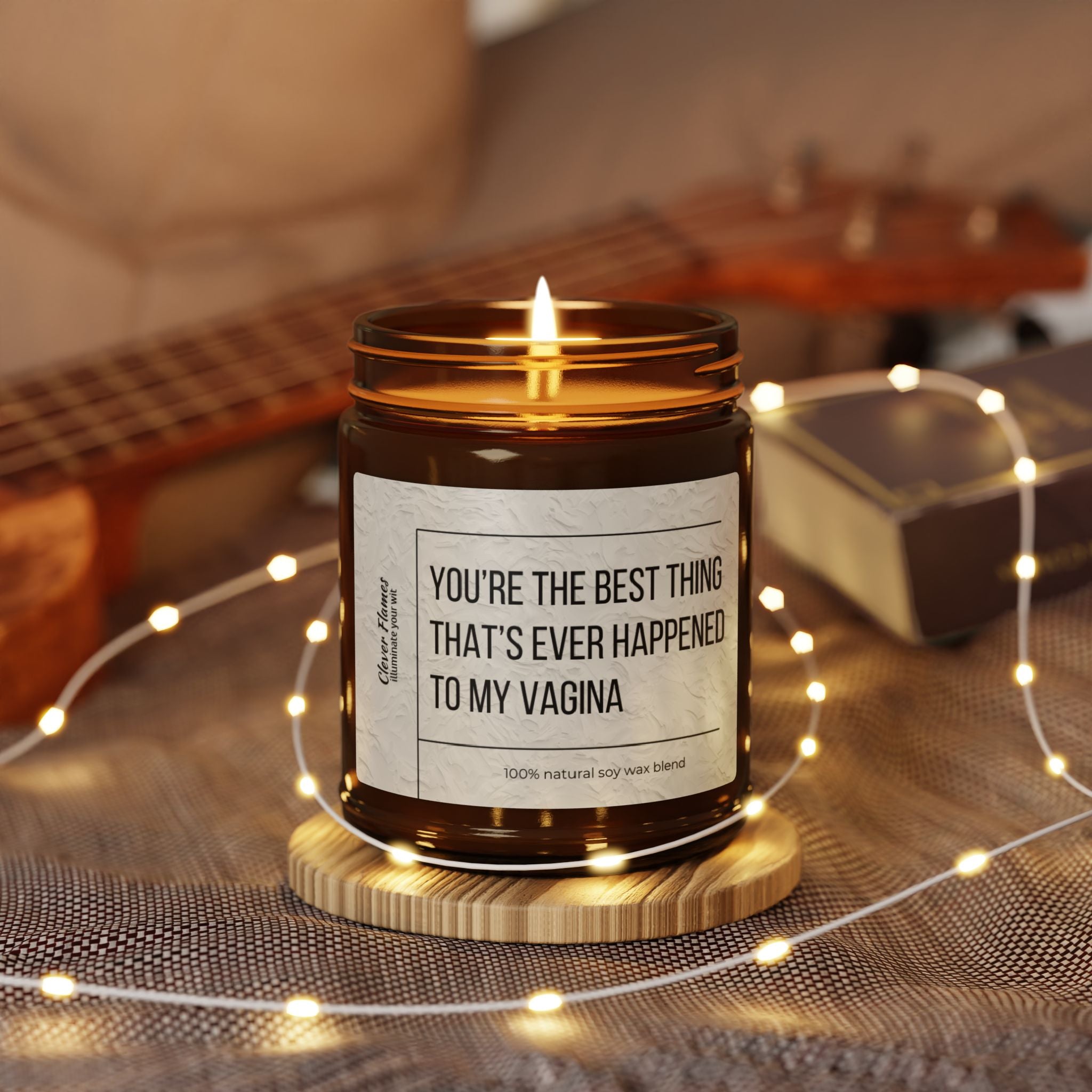 A Printify Best Thing Thats Ever Happened To My Vagina Sex Candle rests on a round coaster, encircled by fairy lights evoking Christmas warmth, with a blurred ukulele in the background.